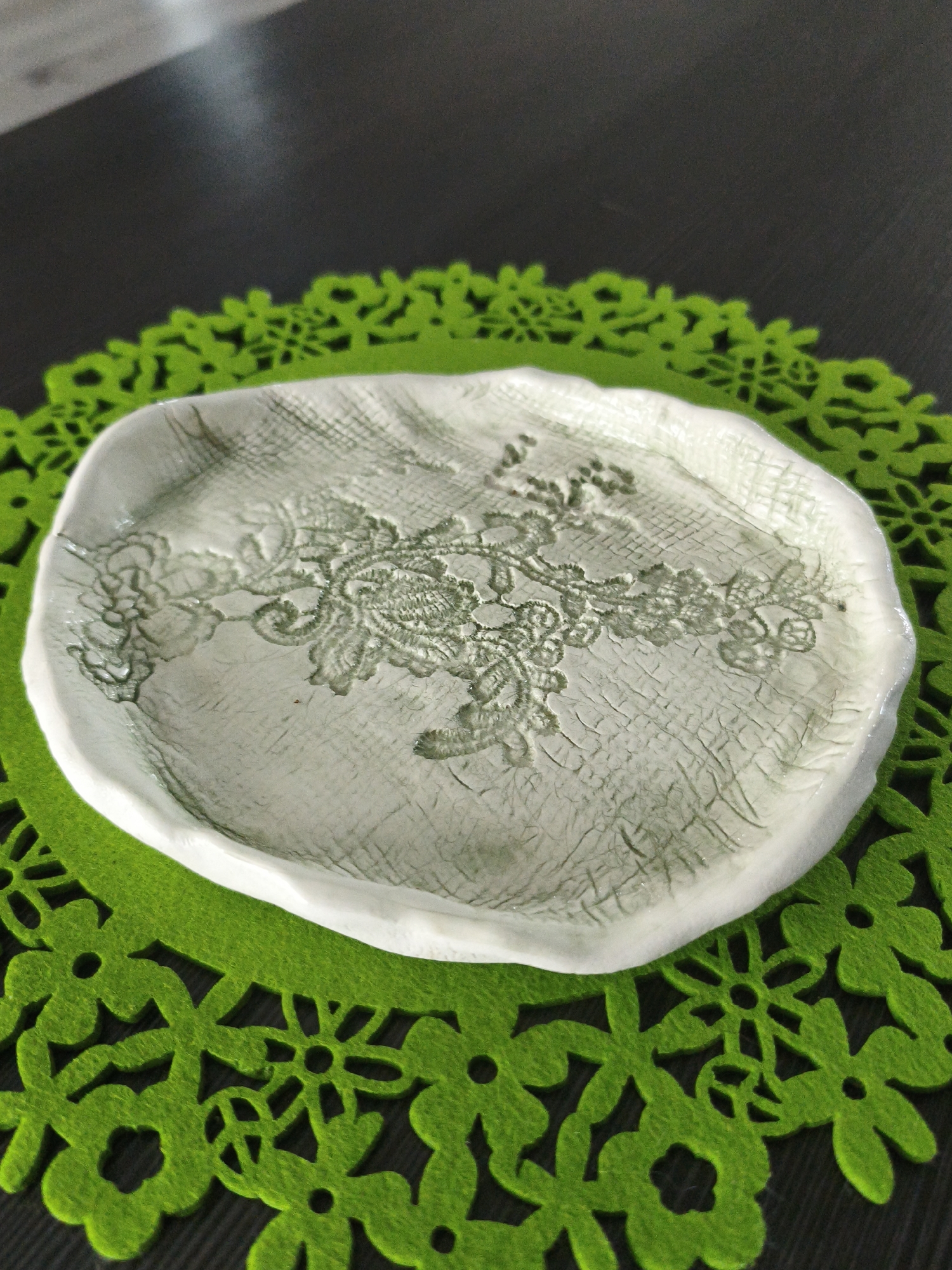 A close-up, high-angle shot shows a small, off-white ceramic dish with a lace pattern pressed into its surface. The dish has an irregular, organic shape with a slightly raised rim. The lace design is in a darker, greenish-grey tone and covers the interior of the dish. The ceramic has a matte finish with visible crackling. The dish rests on a bright green, felt placemat with a cut-out floral pattern. The placemat is on a dark, textured surface, possibly a wooden table. The lighting is soft, creating subtle shadows. There are no people or animals present in the image. The location in the city of Brodi, Latvia, is not discernible from the visual information provided.