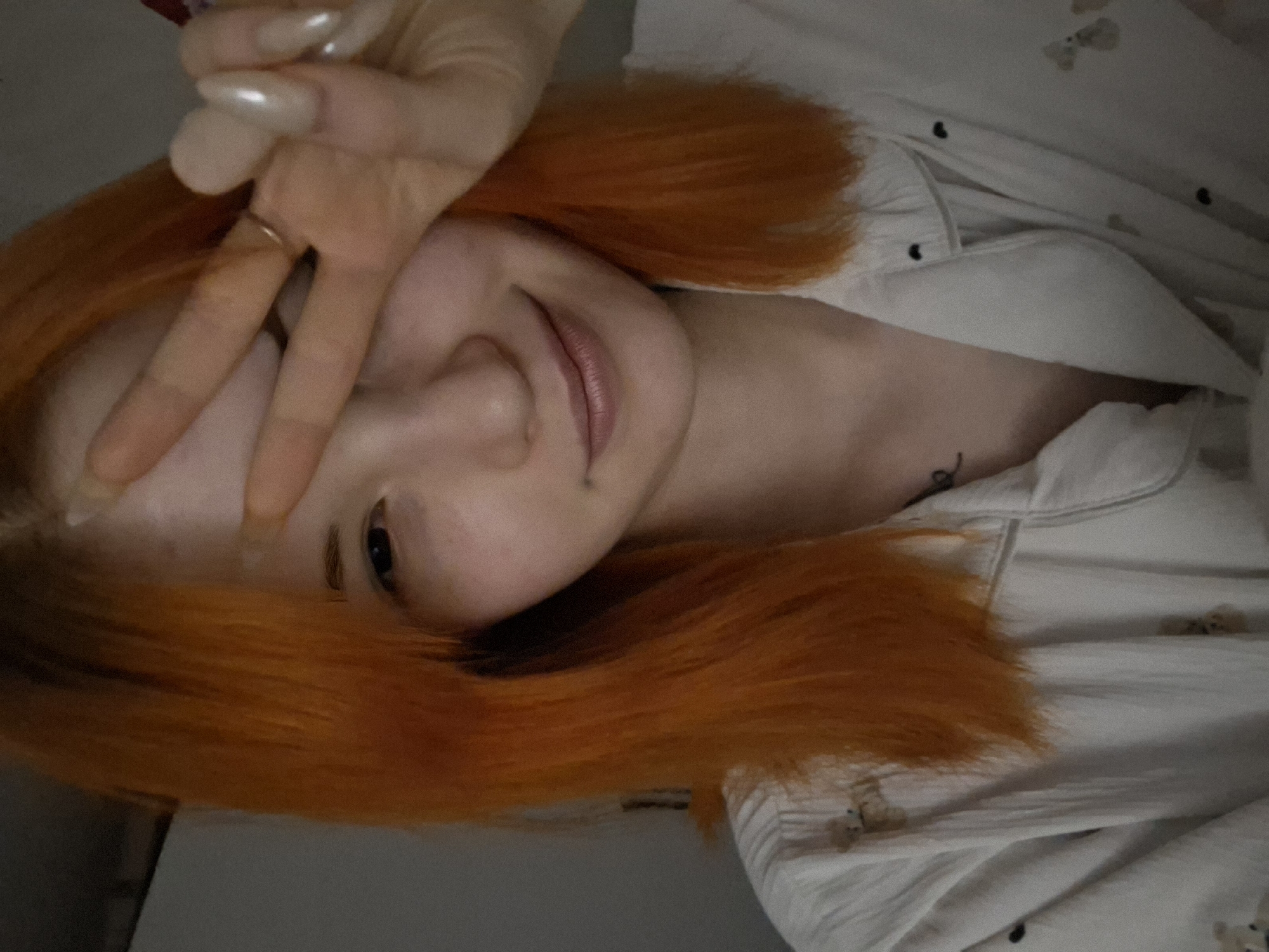 This media file presents a close-up, sideways-oriented portrait of a person, likely a young woman, with vibrant orange hair. She is captured in what appears to be a casual selfie. Her left hand is raised, forming a peace sign (V-sign) with her index and middle fingers, which partially covers her face. She has long, light-colored, possibly pearlescent, artificial nails and a thin, gold-toned ring on her ring finger.
Her visible eye, the left one, is dark and looks directly at the camera, enhanced with what appears to be eyeliner or mascara. Her lips are gently parted in a soft smile, wearing a light-colored lip gloss or lipstick. A small, dark tattoo or mark is visible on her neck or collarbone area. She is dressed in a light-colored, possibly white or cream, pajama-style top that features a faint pattern of small, dark, stylized figures, possibly teddy bears.
The background is softly blurred, indicating an indoor setting, likely a bedroom, given the relaxed attire. The lighting is gentle and diffused, suggesting an intimate personal moment. The overall emotion conveyed is one of relaxation, friendliness, or playfulness, expressed through her soft smile and hand gesture. There is no discernible text within the image. The image itself provides no specific visual cues to its location in Chaikovsky, Russia, beyond the general context.