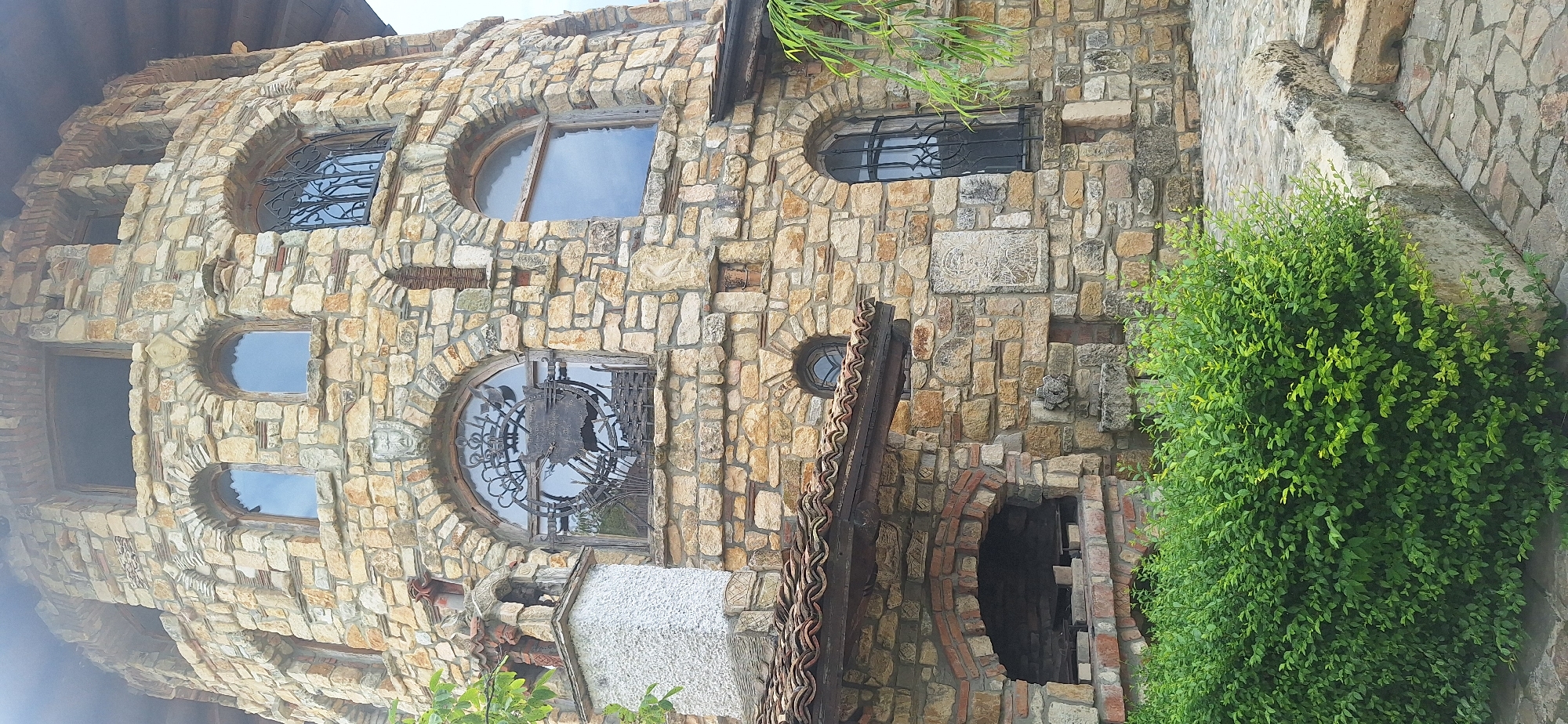 The image shows a stone tower in Kalimantsi, Bulgaria. The tower is made of rough-hewn stones of various sizes and colors, predominantly tan and grey. It features several arched windows, some with decorative ironwork and others with simple wooden frames. Below the tower, there is a covered area with a tiled roof, resembling an outdoor oven or barbecue pit made of brick. In the foreground, a dense green hedge is visible, and a stone-paved walkway leads to a set of stone steps on the right side of the image. The overall scene depicts a rustic, architectural structure in an outdoor setting.