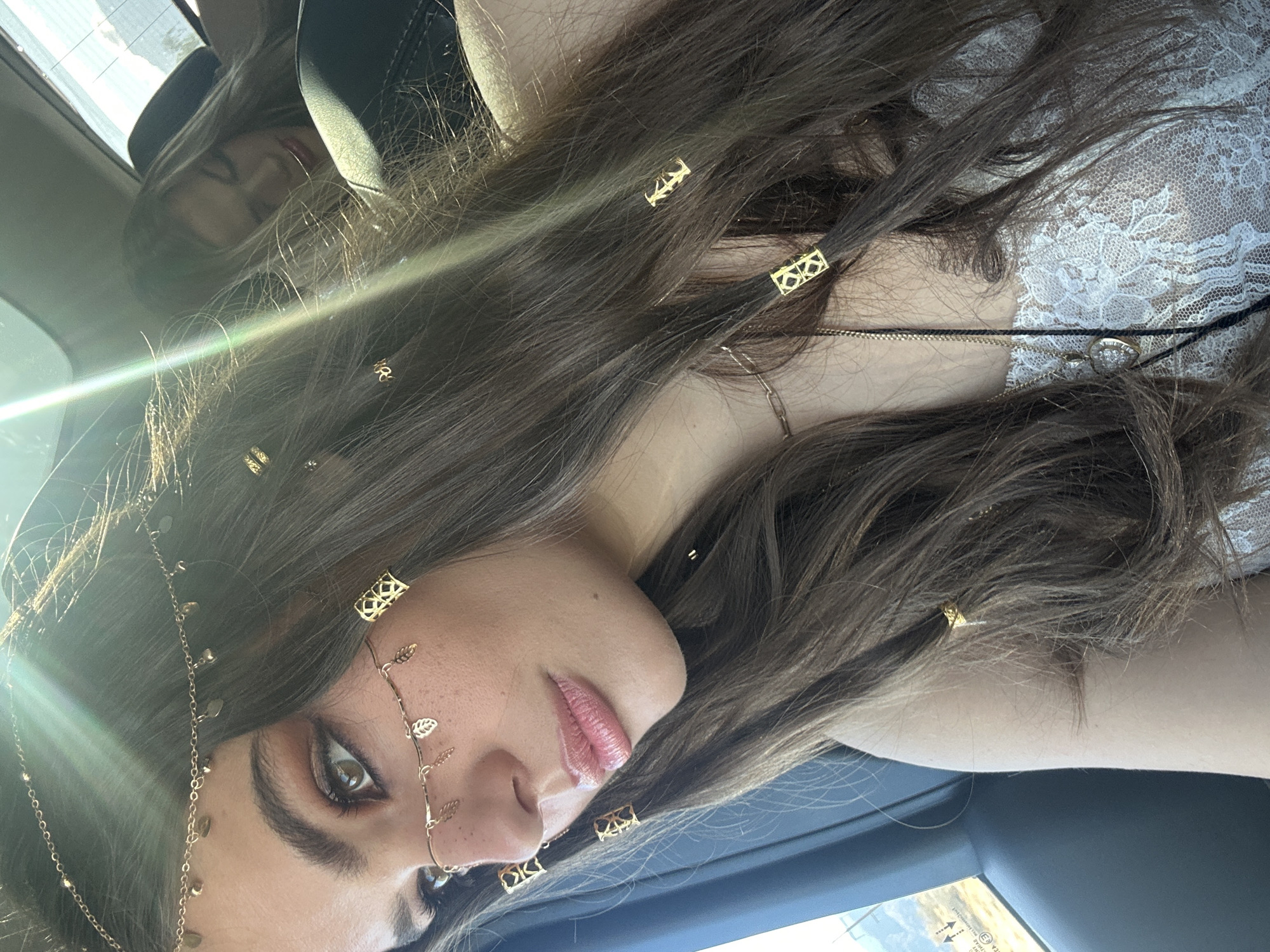 The image shows a woman with long, brown hair adorned with gold-colored hair accessories. Some are rectangular, while others feature leaf-like designs. She is wearing a chain across her forehead and nose. Her skin appears fair, and her lips are glossy. She is in the front seat of a car. A second woman is in the back. The image was taken in Palm Desert, United States of America.