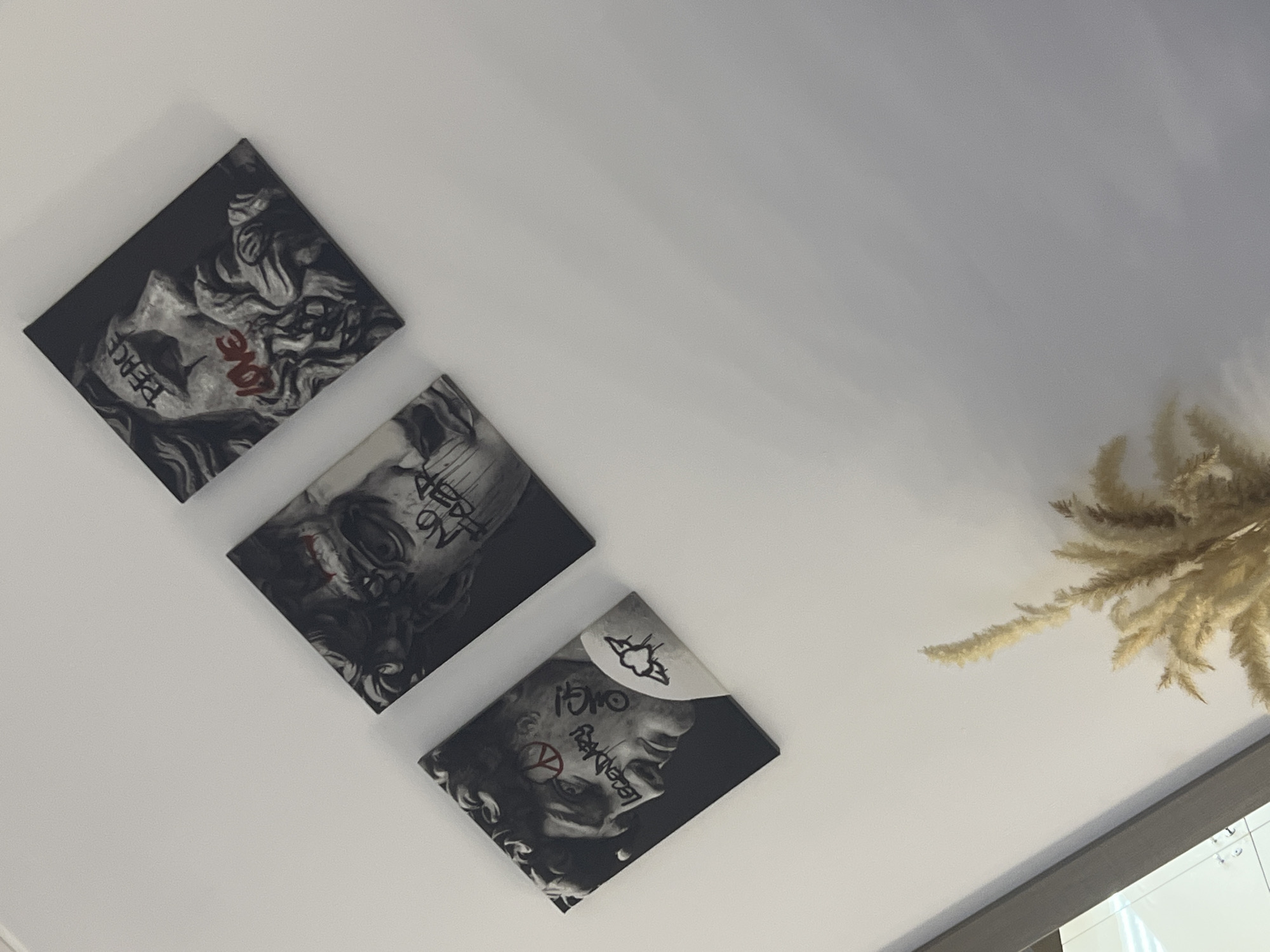 An indoor scene is depicted, featuring a white wall angled slightly upwards from left to right. Three rectangular canvases, oriented diagonally, are mounted on the wall in a staggered arrangement. Each canvas portrays a grayscale stylized image of a classical male bust or sculpture, defaced with graffiti-style text and symbols in red and black.
The leftmost canvas shows a profile of a bust facing right, with a red peace symbol on its forehead, the word "LEGENDARY" in black on the cheek, and "OUCH" in black on the neck. A black outline of a winged heart is visible on the shoulder area.
The middle canvas displays a bust with obscured facial features, partially covered by black text that includes "NO FEAR" and drip effects, along with red markings on the forehead.
The rightmost canvas features a profile of a bust facing left, with the word "PEACE" in black on its forehead and "LOVE" in red with drip effects across its cheek and beard.
In the lower right corner of the frame, a cluster of light beige, feathery ornamental grasses or pampas grass is visible, partially extending upwards from the bottom edge. A dark, reflective surface, possibly a mirror or framed glass, is partially visible along the lower left edge. No individuals are present, and no actions or interactions are depicted. This scene is located within a structure in Krasnoyarsk, Russia.