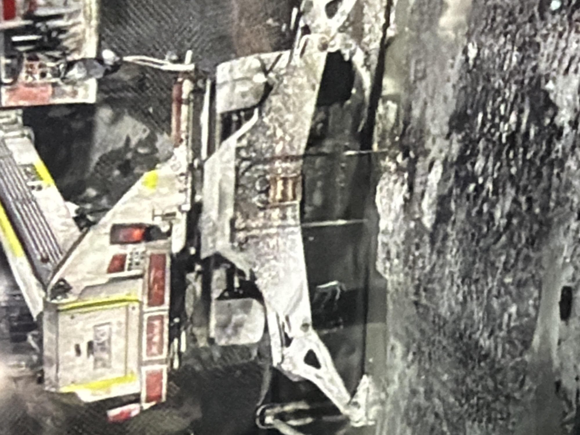 The image shows a construction vehicle, possibly a crane or excavator, in a muddy, industrial-looking environment. The vehicle is predominantly white and grey with yellow and red accents. It appears to be stationary, and there are no people visible in the frame. The ground is covered in dark mud and what looks like debris or rough terrain. The overall impression is one of a work site or an area undergoing construction or demolition.