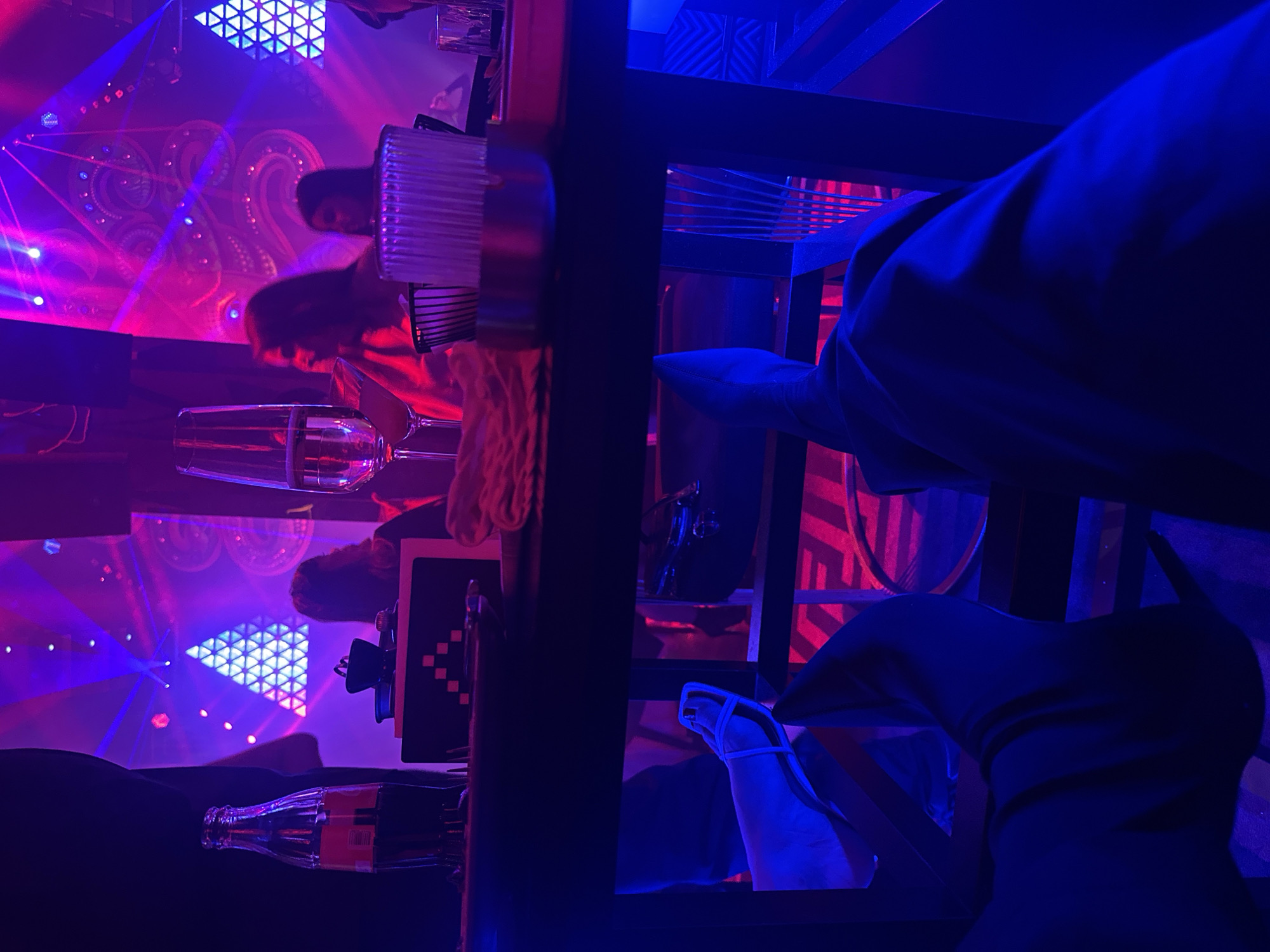 This media file depicts a vibrant, dimly lit indoor scene, characteristic of a nightclub or bar, located in Yerevan, Armenia. The space is bathed in intense neon blue and pink/purple lighting, which casts dramatic shadows and highlights across the room. Streaks of light, resembling laser beams, cut through the hazy atmosphere, contributing to an energetic ambiance.
In the midground, several people are gathered around a table or bar counter. Their upper bodies and faces are partially visible, with some features obscured by the low light and composition. On the table, various drinks are present, including a champagne flute filled with liquid, a Coca-Cola glass bottle with its distinct red label and "Coca-Cola" text visible, and another glass with a ribbed design.
The background showcases impressive decorative lighting, featuring a prominent triangular LED pattern composed of many smaller triangles, emitting bright white and blue light. Swirling circular patterns are also visible on a distant wall, adding to the dynamic visual appeal.
In the foreground, viewed from a low angle, are the legs and feet of a person seated under the table. One leg is clad in a dark, form-fitting boot, while the other foot wears a light-colored, open-toed sandal. The attire and setting suggest a social event taking place at night, focused on entertainment and enjoyment. The overall mood is lively and engaging.