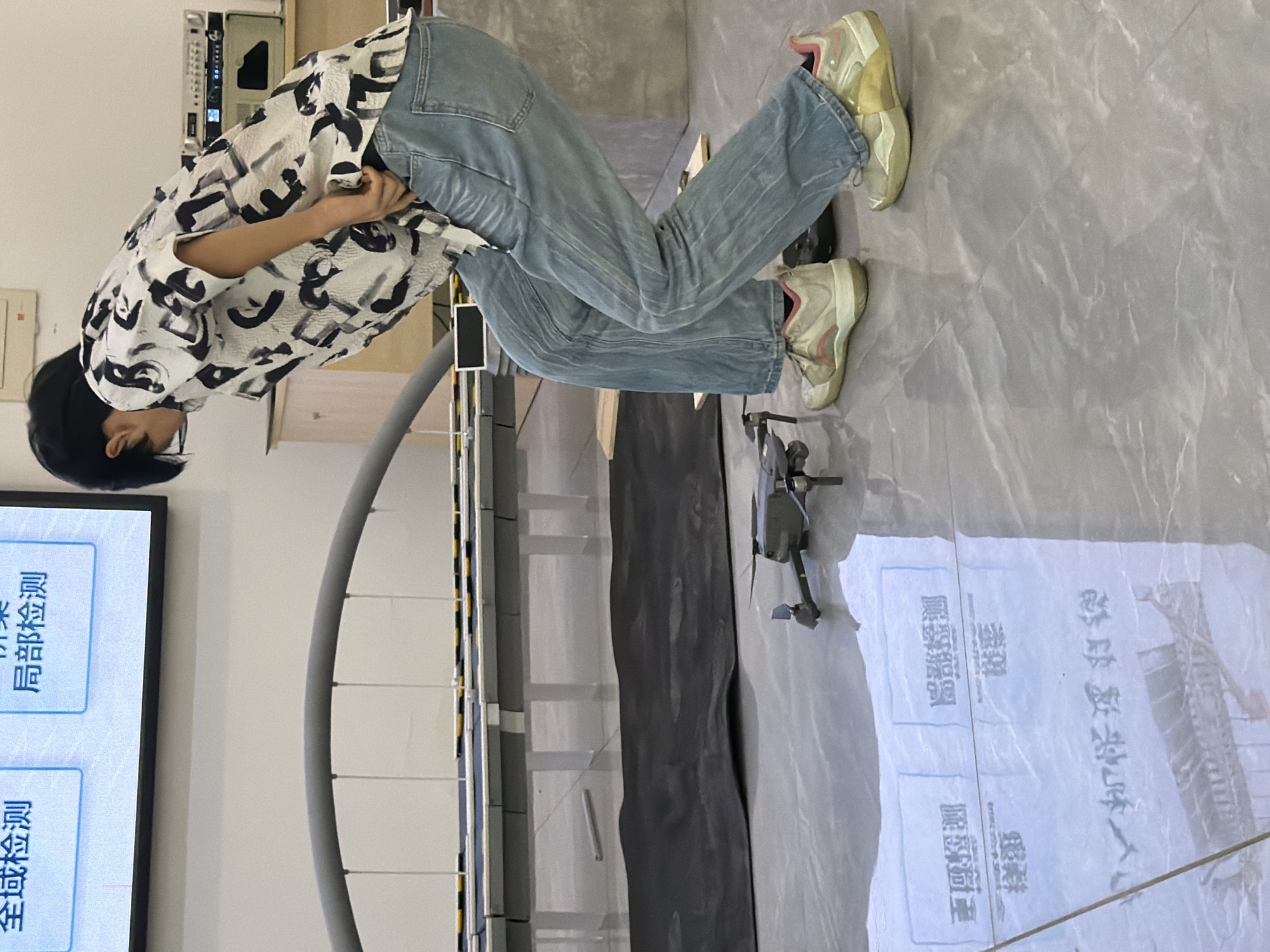 The image depicts a person, identified as a young individual with dark hair, wearing a white short-sleeved shirt with a black abstract pattern and light blue denim jeans. They are wearing athletic shoes, one of which is predominantly white with pink and yellow accents, and the other appears to be white with yellow and pink accents. The individual is in a crouched position, looking downwards and to their left.
In front of the person, on a polished grey tiled floor, is a small, dark-colored drone. The drone is positioned with its propellers visible. The floor is reflective, showing a mirrored image of the drone and the person's lower legs and shoes.
In the background, there is a large screen displaying text in Chinese characters. To the left of the screen, an arched structure made of grey material supported by white vertical elements is visible. Further in the background, on a lighter-colored surface, there are some electronic devices and a wooden cabinet. There is also a dark, rectangular mat on the floor behind the drone.
The scene appears to be indoors, possibly in a laboratory or testing facility, given the presence of the drone, the screen with text, and the general tidiness of the environment. The text on the screen and the reflections on the floor suggest a setting where technological testing or analysis might be taking place. The location is identified as Jiangjin District, Chongqing, China.