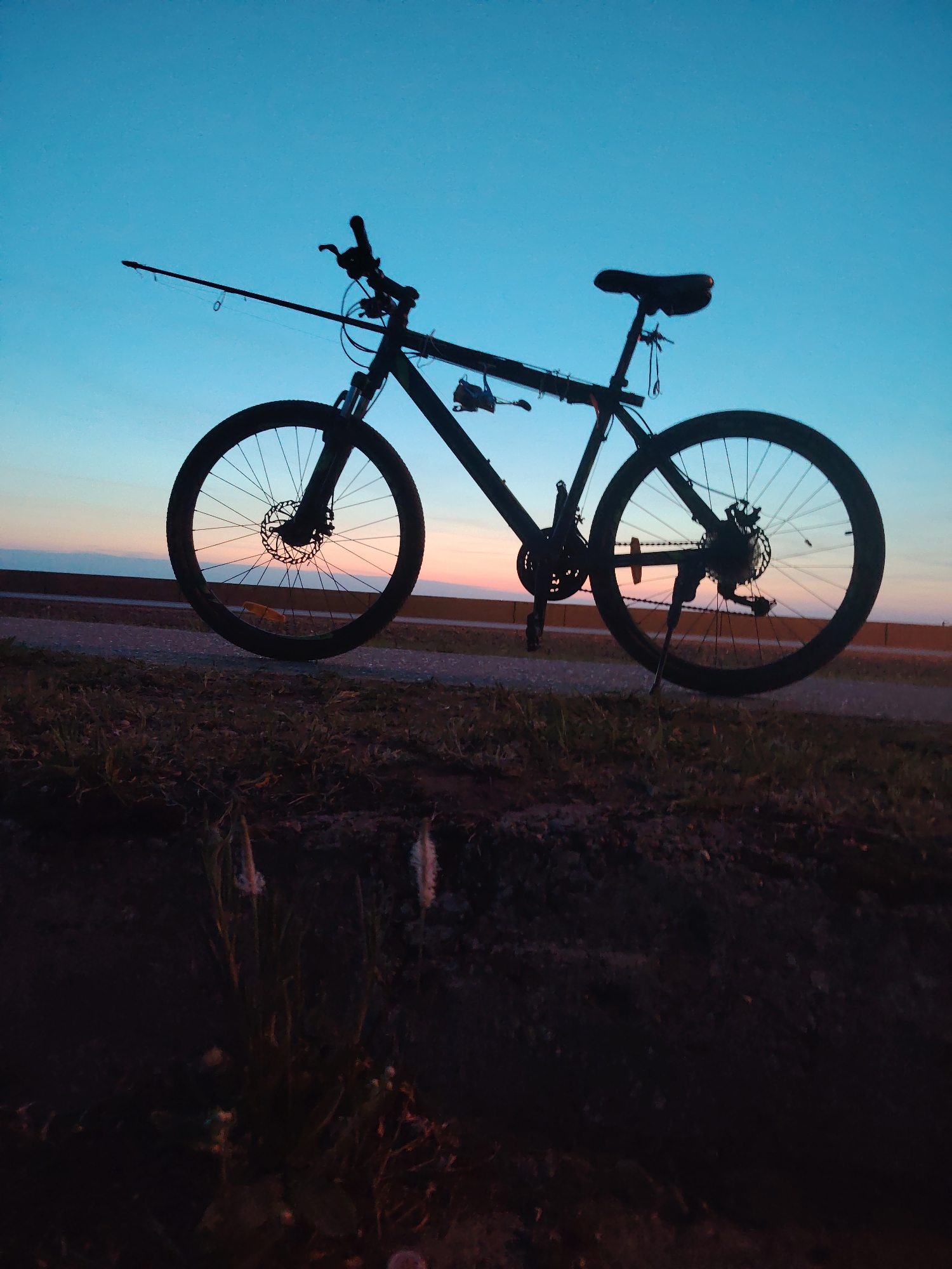 A silhouette of a bicycle, with a fishing rod attached to the frame, stands on a grassy area next to a paved walkway. The background reveals a gradient of colors in the sky, transitioning from orange near the horizon to blue higher up, likely indicating either sunrise or sunset. The location is suspected to be the city of Chaikovsky, Russia. No people are visible. The bike is parked with its kickstand down.
