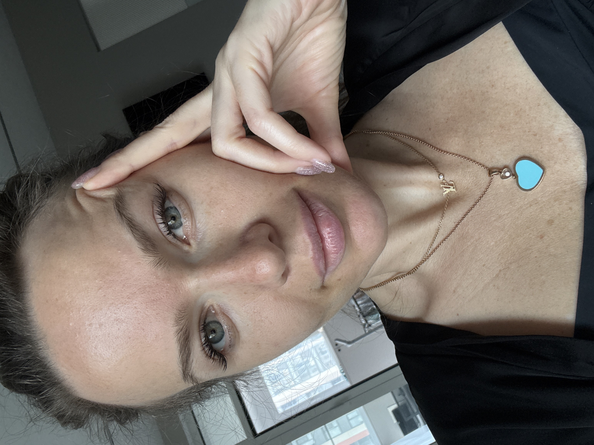 A woman is taking a selfie indoors, likely in an apartment. She is positioned facing the camera and is holding her hand up to her forehead. She has light skin, light-colored eyes, and appears to be wearing little or no makeup. She has a gold necklace with a pendant in the shape of a heart and a letter "K". She is wearing a black garment. A window is visible in the background showing what appears to be an urban cityscape, possibly Dolgoprudny, Russia, as indicated by the prompt. The cityscape includes modern buildings and possibly a crane. A window blind and some type of light fixture are also visible in the top-left corner.