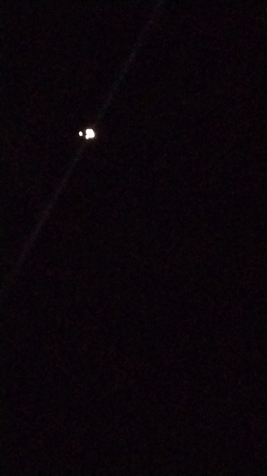 The image depicts a dark, nighttime scene, likely taken outdoors. In the upper left quadrant, there are two bright, indistinct white or light yellow celestial bodies, one slightly larger and more diffuse than the other, suggesting they might be stars or the moon. A faint, diagonal light streak or reflection is visible extending from the upper left corner towards the celestial bodies. The rest of the image is predominantly black, indicating a lack of ambient light and a likely high contrast photographic setting. There are no discernible man-made structures or natural features of Madduru, India, visible in the image.