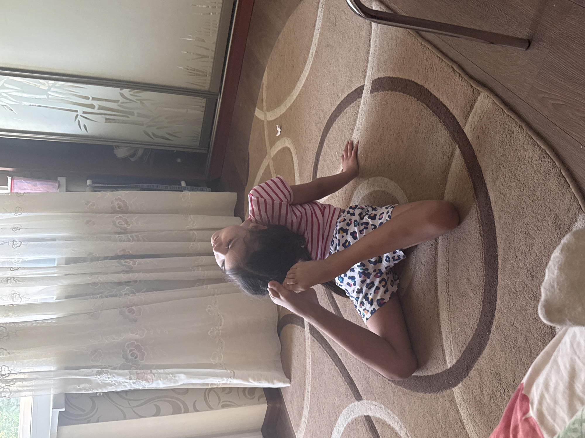 A young person, possibly a child, with dark hair, lies on their back on a light brown patterned carpet, performing a yoga or gymnastic pose. They are wearing a pink and white striped short-sleeved top and light-colored shorts with a dark animal print. Their legs are bent, and their hands are grasping their feet, arching their back and tilting their head upwards. The carpet features a pattern of concentric light and dark brown circles and curved lines. In the background to the left, a window is covered by sheer white curtains with embroidered floral patterns, partially revealing light grey patterned wallpaper. To the right, a tall wooden wardrobe or cabinet with sliding doors is visible; one door features a frosted or etched design of bamboo stalks. A small white crumpled object rests on the carpet near the child's right side. A metallic furniture leg is visible on the far right. A blurry red and white patterned fabric is present in the lower-left foreground. The room is brightly lit, with natural light entering from the left.
Location: Unknown, Unknown.