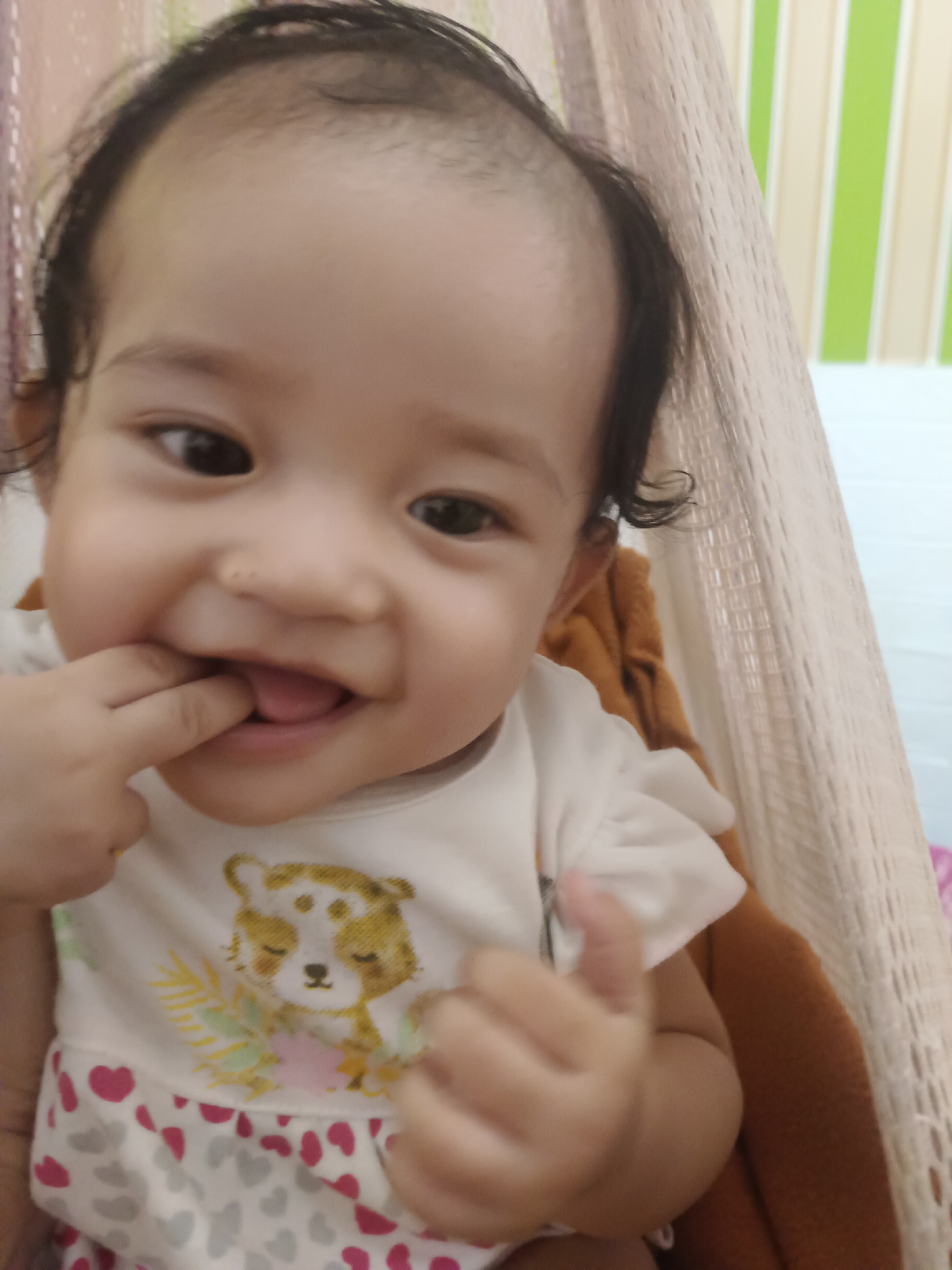 The image shows a close-up of a baby's face and upper body. The baby is wearing a white shirt with a print of a cartoon animal and hearts. The baby has dark hair and is looking at the camera with a smile, with one hand in their mouth. The background consists of a light-colored hammock and a wall with vertical stripes. The scene appears to be indoors. There is no indication of the city of Kawua, Indonesia, in this image.