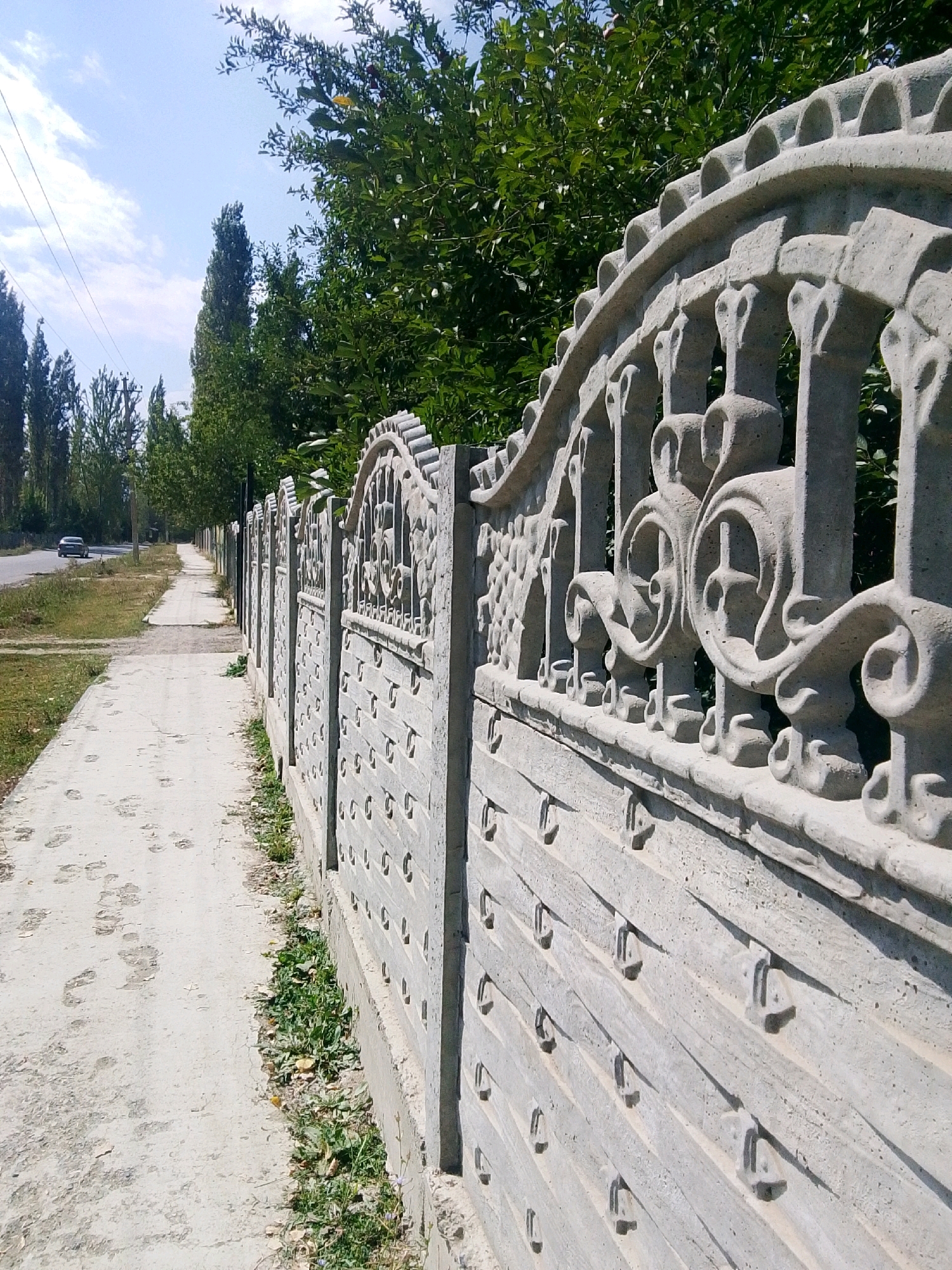 The image shows a concrete fence with decorative elements alongside a sidewalk. The fence is light gray and features a patterned design with circular and curved motifs on its upper portion. Below the decorative elements, the fence has a textured surface with small, raised rectangular protrusions arranged in rows. The sidewalk is paved and appears somewhat worn, with visible impressions that could be footprints or tire marks. To the left of the sidewalk is a grassy area with some fallen leaves and weeds. Beyond the fence and grass, a road is visible with a single car driving away in the distance. A line of tall, slender trees with dense green foliage borders the road and the area beyond. The sky is bright blue with scattered white clouds, suggesting it is daytime and likely sunny. The overall scene depicts a street view in what is described as the city of Kara-Jygach, Kyrgyzstan, characterized by residential or public landscaping.