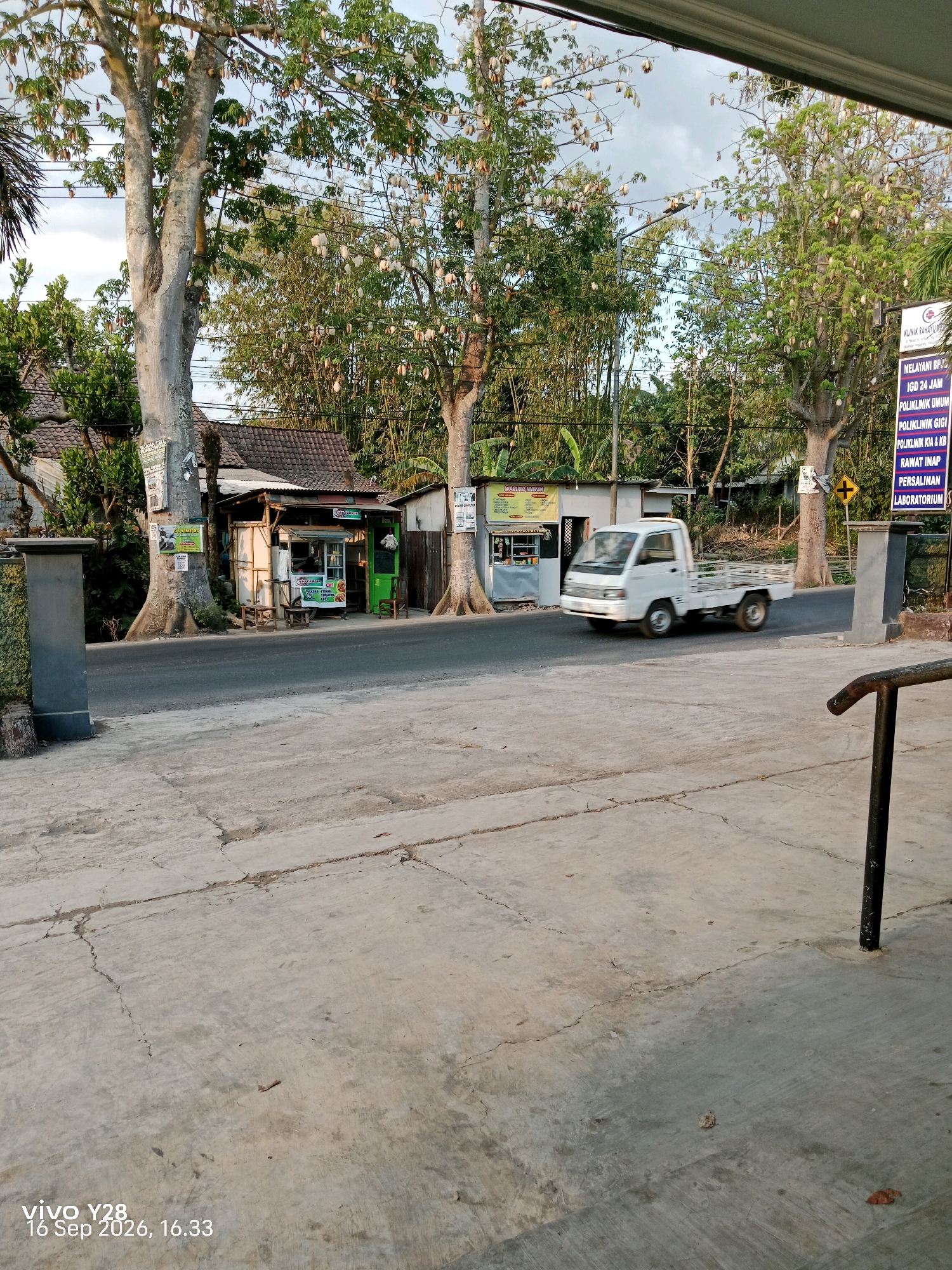The image shows a street scene in Panggungan, Indonesia, featuring a white truck in motion on a paved road. The road is lined with trees, small buildings that appear to be shops, and concrete sidewalks. One of the buildings has a sign that reads "KLINIK RAHAYU" and lists various medical services. Another small shop displays the text "MAGUNG MAKAN". The sky above is overcast. The foreground of the image is a cracked concrete surface. The timestamp "vivo Y28, 16 Sep 2026, 16.33" is visible in the bottom left corner.