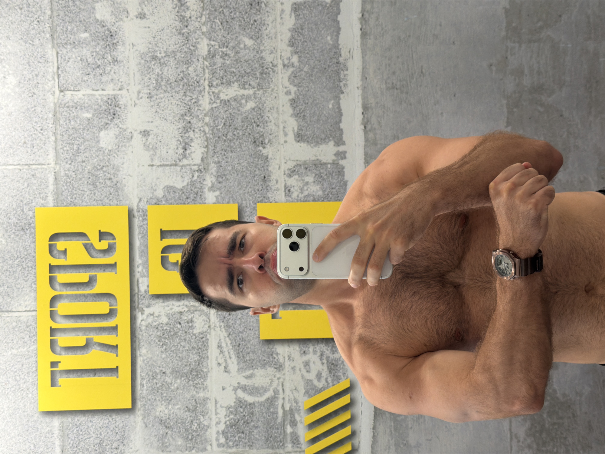 A shirtless man with dark hair, a muscular build, and a watch on his left wrist stands in front of a grey brick wall in what appears to be a gym. He is holding a white smartphone in his right hand, taking a selfie. Yellow signage with cut-out letters spelling "SPORT" is visible on the wall behind him. The overall environment suggests a fitness facility in Tel-Aviv, Israel.
