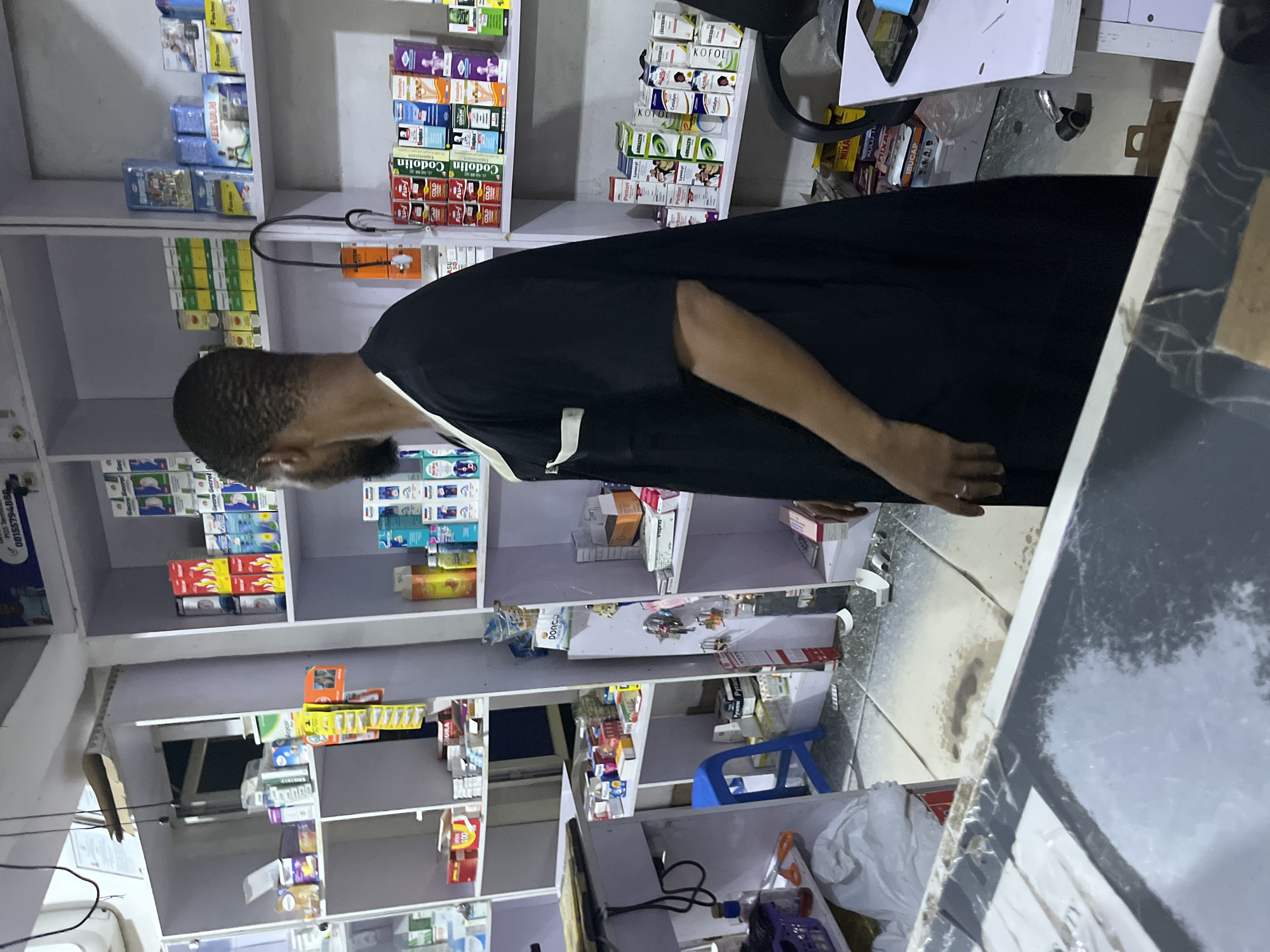 Here is a description of the image:
The image shows the interior of a pharmacy, likely located in Abuja, Nigeria, based on the products and layout. A man with dark hair and a beard, wearing a black garment with a white trim on the collar, is standing, seemingly looking at the shelves stocked with various medications and pharmaceutical products. The shelves are white, and items are arranged in boxes and bottles. There is a counter with a dark surface partially visible in the foreground. The floor is tiled, and several items are scattered around it.
