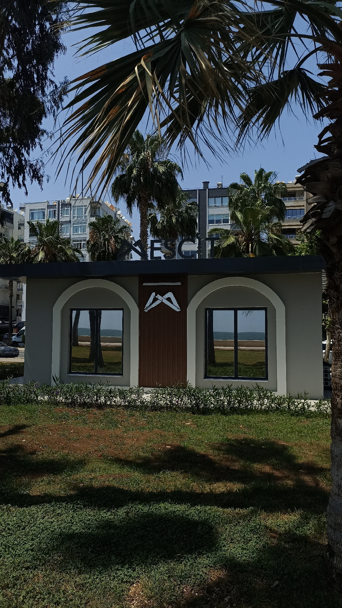 The image captures a bright, sunny scene featuring a small, modern building set in a green area, likely in a coastal or resort setting given the surrounding elements. The building itself is light grey with dark grey arched frames around two prominent windows and a central wooden panel. A white, stylized emblem resembling an "M" or two inverted "V" shapes is affixed to the wooden panel. Above the building, a dark sign bears the white letters "RESORT", with the first 'R' being stylized.
The windows of the building reflect the bright blue sky and what appears to be the sea or a large body of water on the horizon, along with reflections of palm trees. These reflections indicate a clear, expansive view from the building's location.
In the foreground, lush green grass covers the ground, interspersed with some patches of drier earth. Strong, dark shadows from surrounding palm trees are cast across the grass, highlighting the bright sunlight of the day.
The background reveals several multi-story residential buildings and numerous palm trees, contributing to the typical Mediterranean or resort town aesthetic. The sky is clear and bright blue. No people or specific activities are visible, presenting a serene, static view of the location. The overall impression is one of a well-maintained, sunny vacation spot in Yenişehir, Turkey.