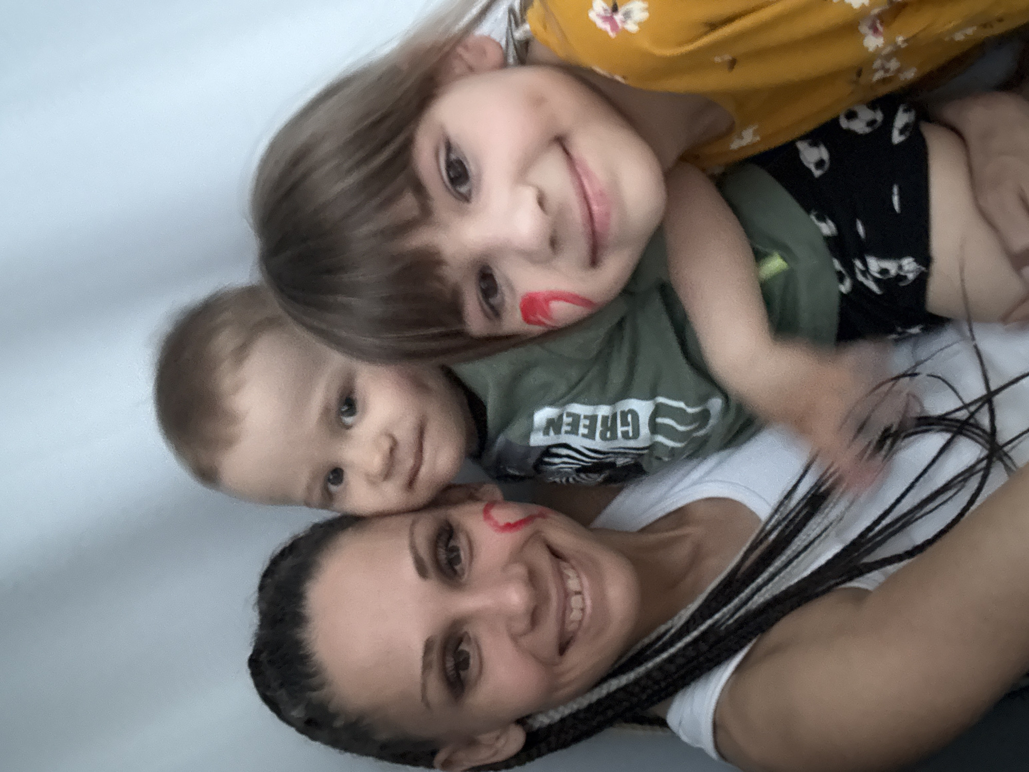 This is an intimately framed, playful selfie featuring three individuals, likely a mother and her two children, stacked on top of each other while lying down. The scene appears to be set indoors in Rusanovo, Russia, against a plain, light-colored background.
At the front of the stack, closest to the camera, is an adult woman. She has dark, braided hair and a warm, broad smile, showing her teeth. A distinct red, heart-shaped mark is playfully drawn on her left cheek. She is wearing a simple white t-shirt.
Behind her, resting on her, is an older child, a girl with light brown hair and bangs. She is also smiling brightly towards the camera, and like the woman, has a red mark on her cheek. She is dressed in a yellow top adorned with white floral patterns, worn over a green long-sleeved shirt, and black shorts featuring white, skull-like or panda-like designs.
At the very top, partially visible and resting beside the older child, is a younger child, a baby boy, with light brown hair. He looks towards the camera with a gentle, soft smile. He is wearing a green t-shirt that clearly displays the word "GREEN" in bold white letters, accompanied by white stripes.
The image captures a joyful and tender family moment, filled with happiness and affection, likely a casual interaction or playtime. The red markings on the cheeks suggest a shared playful activity such as face painting. The overall mood is cheerful and content.