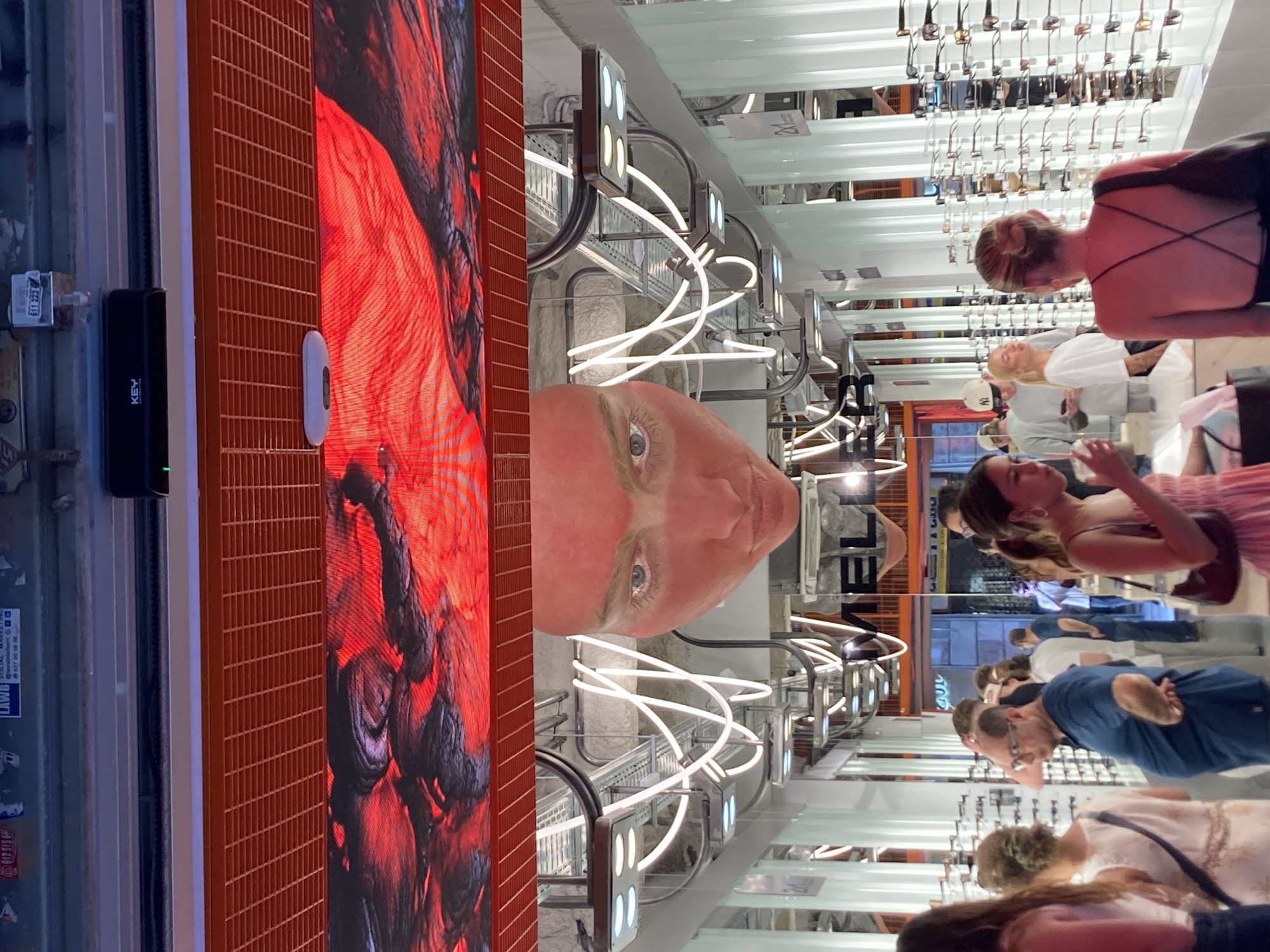 This is a photograph taken at an optical store in Barcelona, Spain. The image shows several people browsing sunglasses displayed on racks. The store appears to be well-lit and modern, with a large LED screen displaying a close-up of a person's eye. The people in the image are casually dressed and seem to be engaged in shopping. Some are looking at the sunglasses, while others are interacting with each other. The overall atmosphere is one of a typical retail environment. Visible text includes brand names, pricing information, and what appears to be promotional material. The time of day is unclear, but the indoor lighting suggests it could be daytime or early evening.