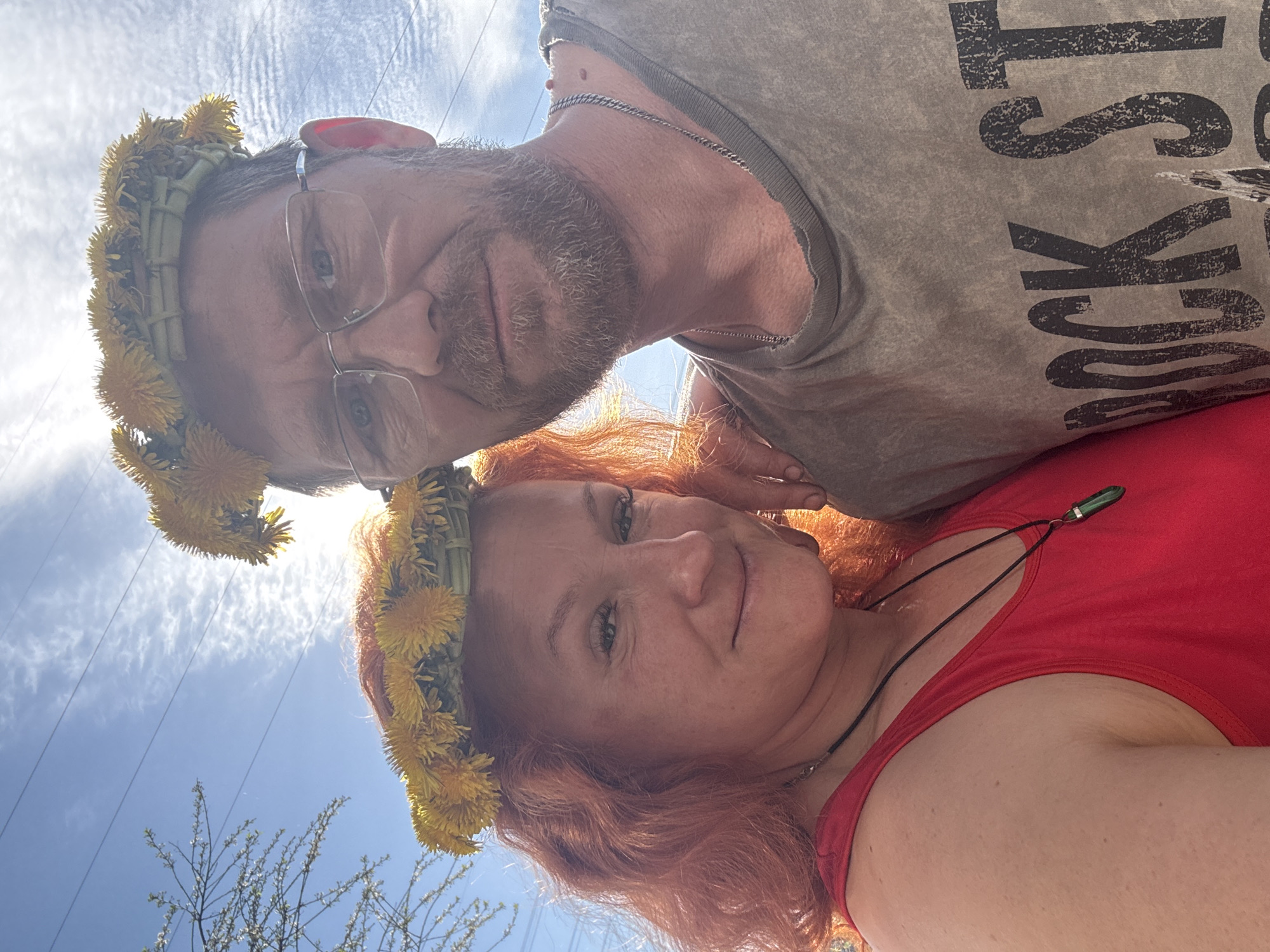 A selfie taken from a low angle features two individuals wearing dandelion crowns. The man, positioned above and to the left, has short brown hair, a beard, and glasses. He is wearing a grey shirt with black lettering that appears to read "ROCK ST." The woman, positioned below and to the right, has red hair and is wearing a red tank top with a necklace. The background shows a clear blue sky with streaky white clouds. Power lines and tree branches are visible in the lower left corner. The location is likely outdoors in daylight. There is no information to indicate this image was taken in the city of Chaikovsky, Russia.