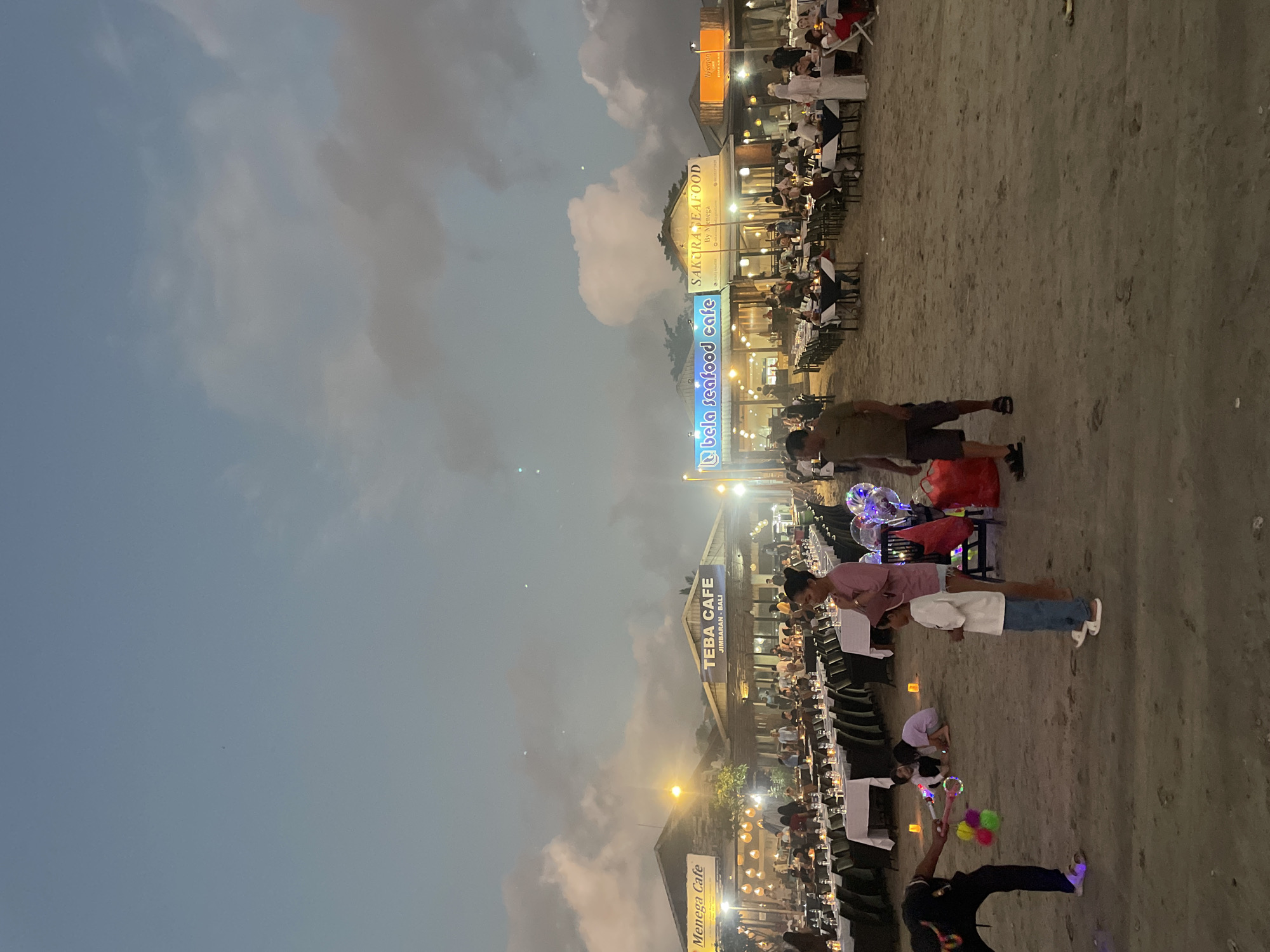 The image depicts a lively beachside dining scene at dusk. Several restaurants are set up on the sand, with long rows of tables covered in white tablecloths and lit by numerous small candles. People are seated at the tables, enjoying their meals. In the foreground, a man is playing with children, blowing bubbles. The sky is a dusky blue with scattered clouds, and lights from the restaurants illuminate the scene. The overall impression is one of a festive and communal dining experience by the sea.