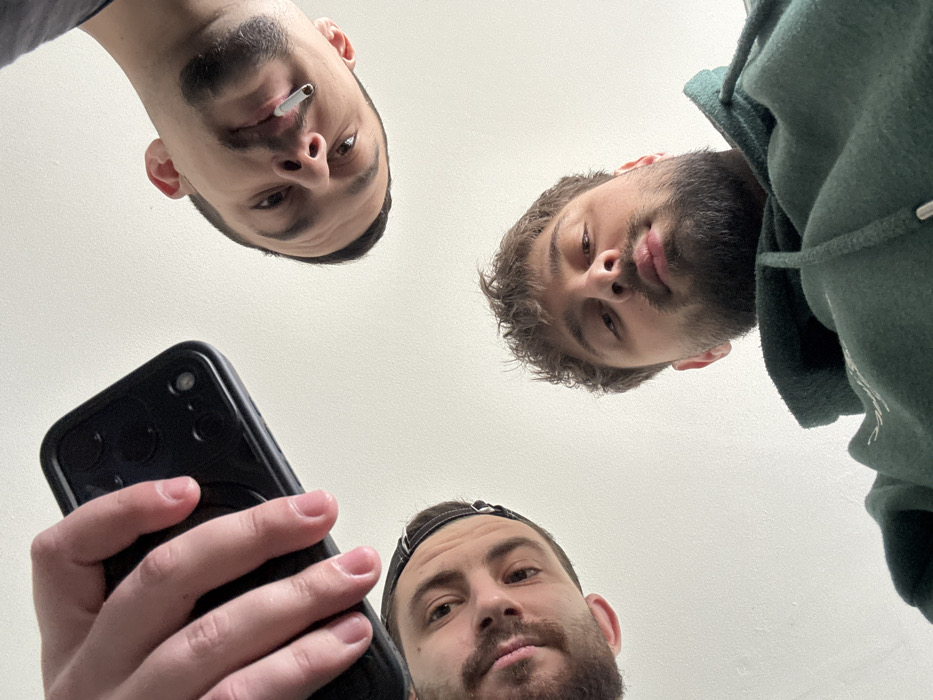 The image displays three adult males looking down towards the camera from a low angle, consistent with an indoor setting in Varna, Bulgaria. In the upper left quadrant, a hand holds a black smartphone with visible camera lenses. To the left, a male with a beard and a dark cap is partially visible. In the upper right, another male with a beard and short hair, wearing a dark shirt, has an unlit cigarette positioned in his mouth. At the bottom center, a third male with a beard and curly hair wears a dark green hooded sweatshirt. The background consists of a plain, light-colored surface, likely a ceiling or wall, providing no specific environmental identifiers beyond its simple, unadorned nature.