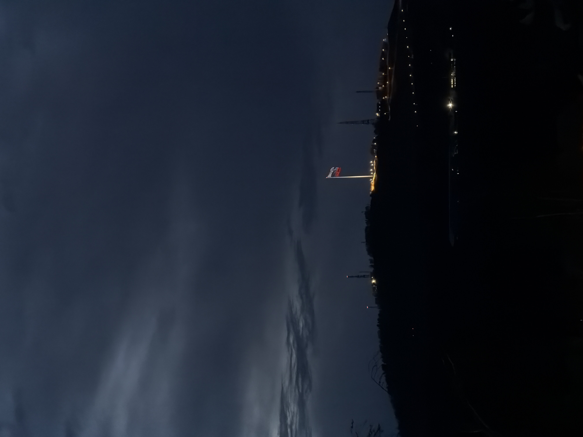 The image captures a night scene in Krasnoyarsk, Russia. In the center of the frame is a flag of Russia flying from a tall flagpole, illuminated by lights. The sky is dark with wisps of clouds near the horizon. The right side of the image shows a dark silhouette of trees and the faint outline of a building, dotted with lights. In the distance, faint light emanates from an industrial area with towers and various structures.
