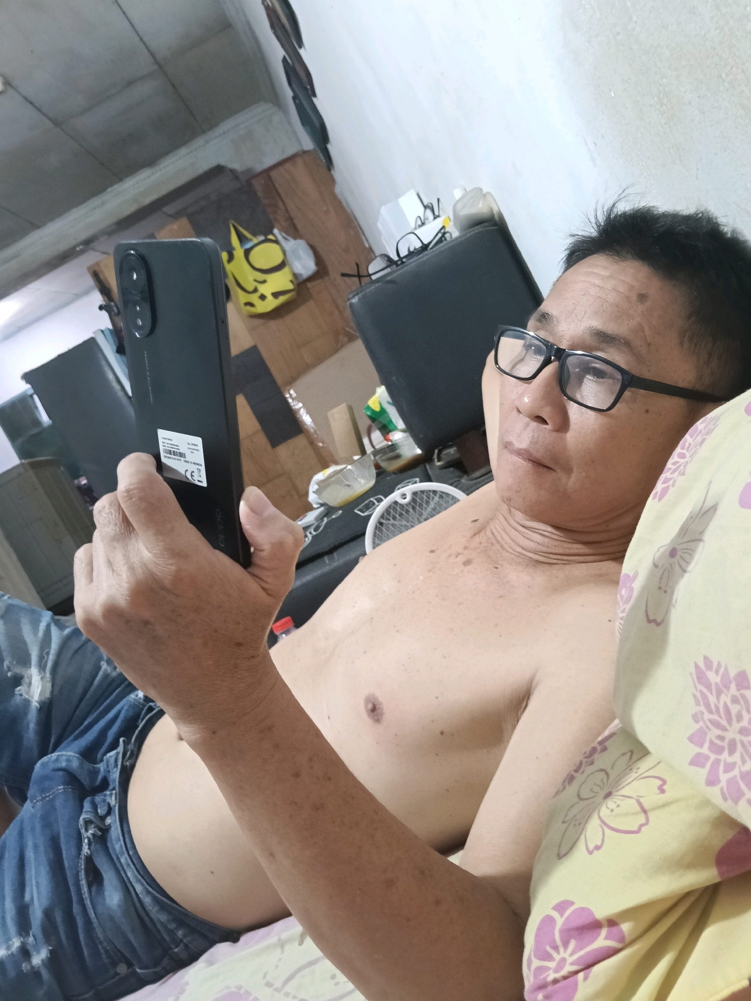 The image shows a man lying on a bed and holding a black smartphone. He is wearing blue jeans and is shirtless. He is wearing black-rimmed glasses. The room appears to be a bedroom with a ceiling fan visible in the background. The walls are light-colored and there are some items on shelves in the background, including a yellow bag with a face. The man is looking at his phone. The bed has a yellow and purple floral patterned pillow. The location is identified as Jatimulya, Indonesia.