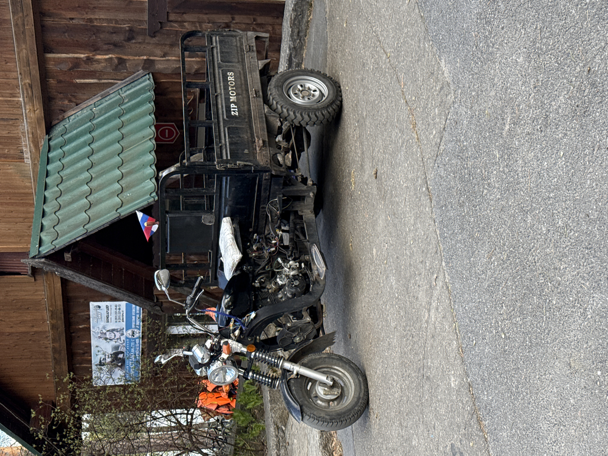 The image captures a ZIP Motors utility vehicle, a three-wheeled vehicle, parked on a paved road alongside a wooden building. The building has an A-frame design with a green corrugated roof extension. Attached to the building is an advertisement in Russian for "BOMBARDIRO", along with phone numbers and an address (Dovatora, 17). The vehicle appears to be in a state of disrepair, with exposed engine components. Based on the OCR text and visual cues, it can be inferred that the location is likely Zolotoi Plyazh, Russia.