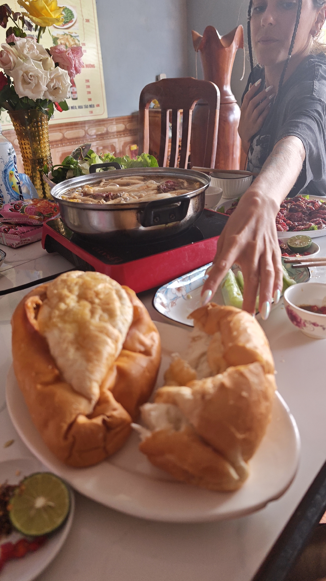 A table is laden with food in what appears to be a restaurant setting in Púng Luông, Vietnam. Dominating the foreground are two large pieces of bread on a white plate. A hot pot with broth and ingredients sits on a red portable induction cooktop. Various dishes and condiments surround the hot pot: leafy greens, a plate with red meat, small bowls with dipping sauces, lime wedges, and cucumbers. A woman reaches for food with her hand. Behind the woman, there is a vase with roses and a decorative wooden object. A menu or sign is partly visible on the wall.