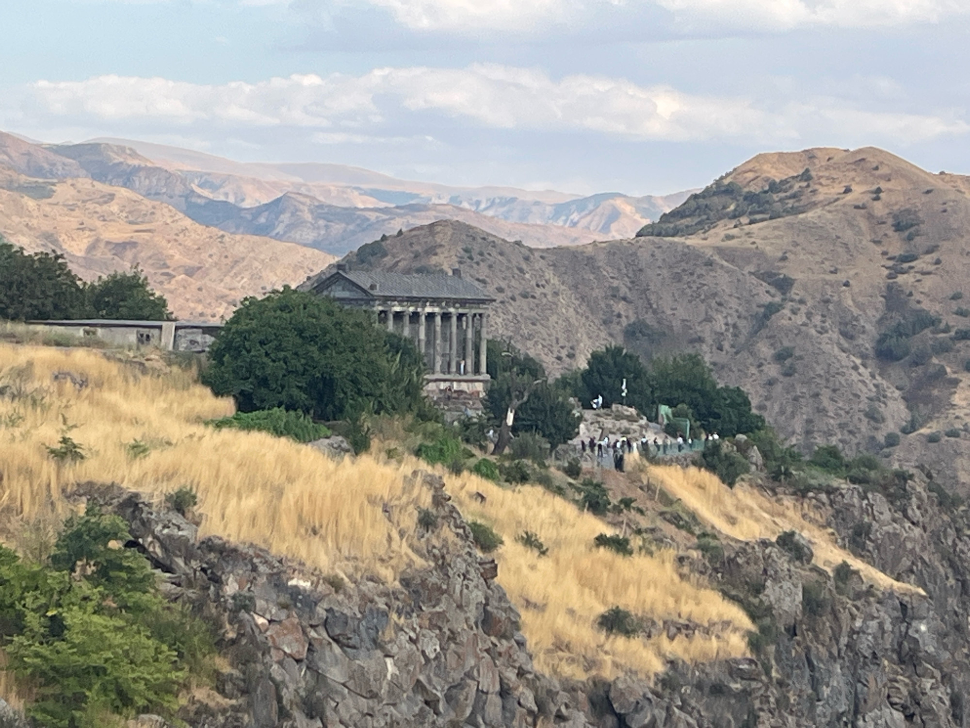 The image depicts the Garni Temple, an ancient Greco-Roman colonnaded temple in Armenia, situated on a rocky cliff overlooking a mountain landscape. The temple, with its prominent columns, stands on a grassy slope dotted with trees. In the foreground, a steep, rocky cliff face is visible, covered in dry, yellow grass. In the middle ground, a group of people are gathered near the temple, some appearing to be tourists. The background consists of arid, mountainous terrain with layers of hills and valleys, under a hazy, overcast sky.