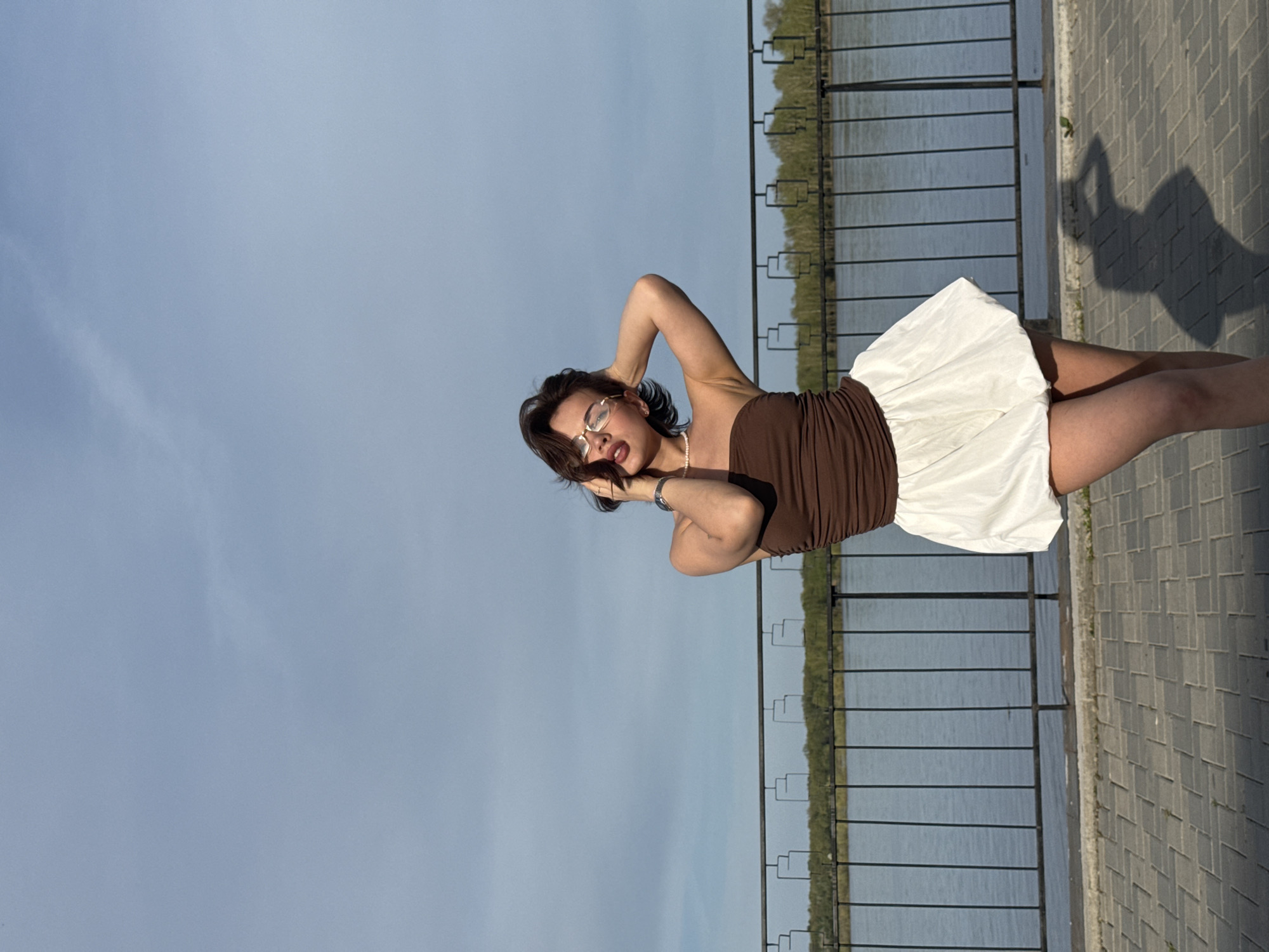 A woman is posing near a railing with a body of water in the background, likely on the banks of the Kama River in Sarapul, Russia. The woman wears a brown top and a white skirt. The area around her appears to be paved. Her shadow is visible on the pavement.