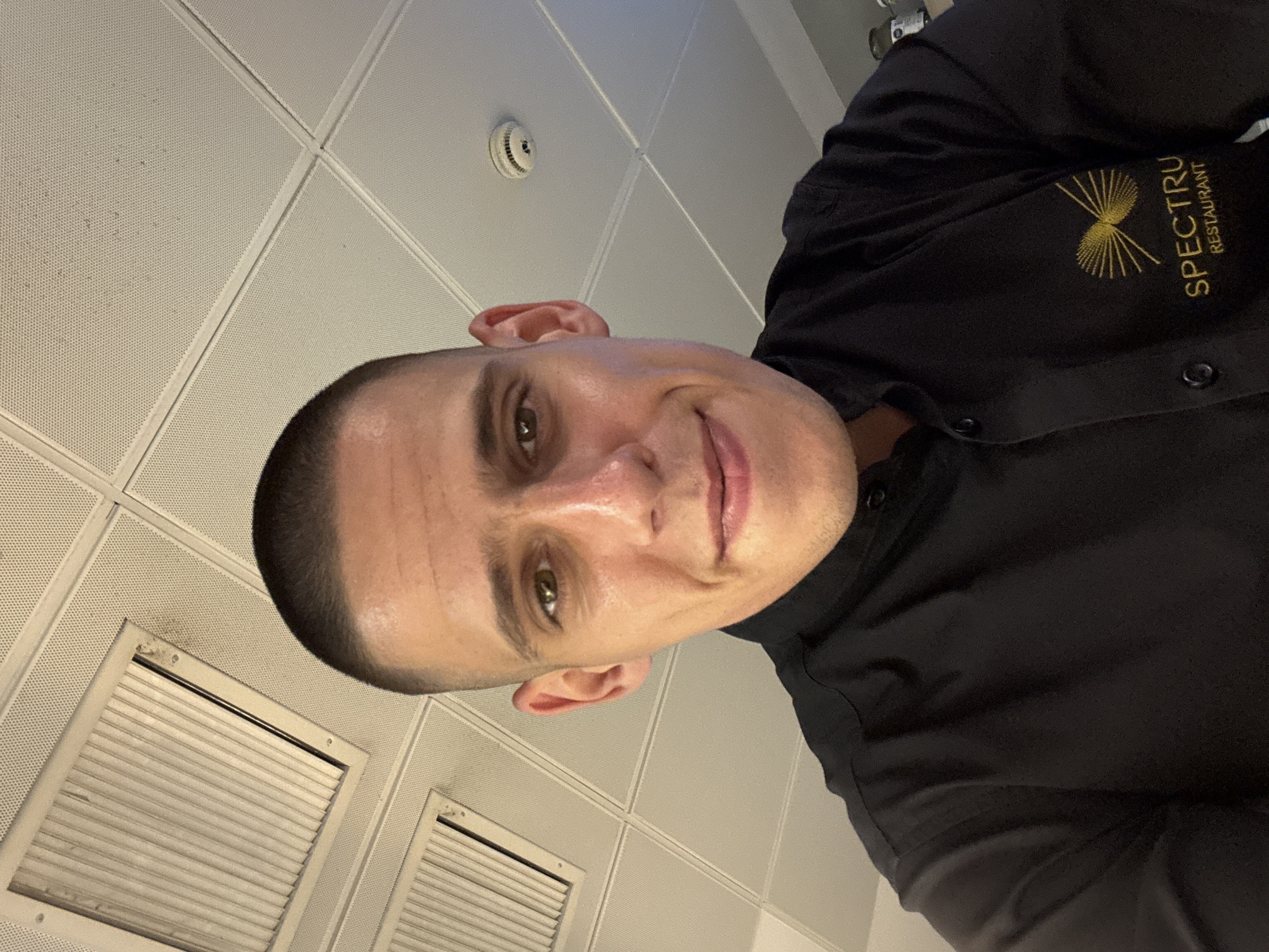 A man with a buzz cut and brown eyes looks up at the camera, smiling slightly. He is wearing a black collared shirt with a gold embroidered logo on the left chest that reads "SPECTRU RESTAURANT". The background is a white drop ceiling with visible light fixtures and what appears to be a smoke detector. The image was taken indoors, likely in an office or commercial establishment, in Sofia, Bulgaria.