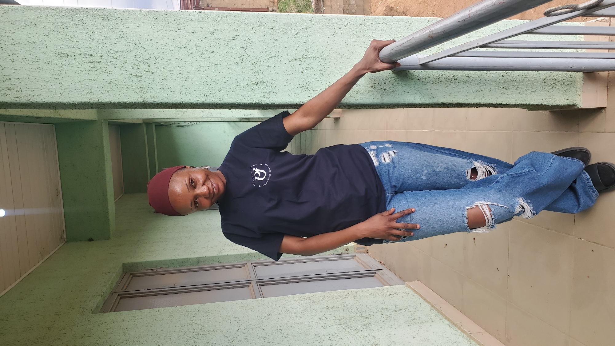 A woman stands at a building in Bukuru, Nigeria. She holds onto a metal railing, wearing a maroon head covering, black t-shirt with a white logo, ripped blue jeans, and black shoes. She stands on a tiled floor adjacent to a light green wall and window.