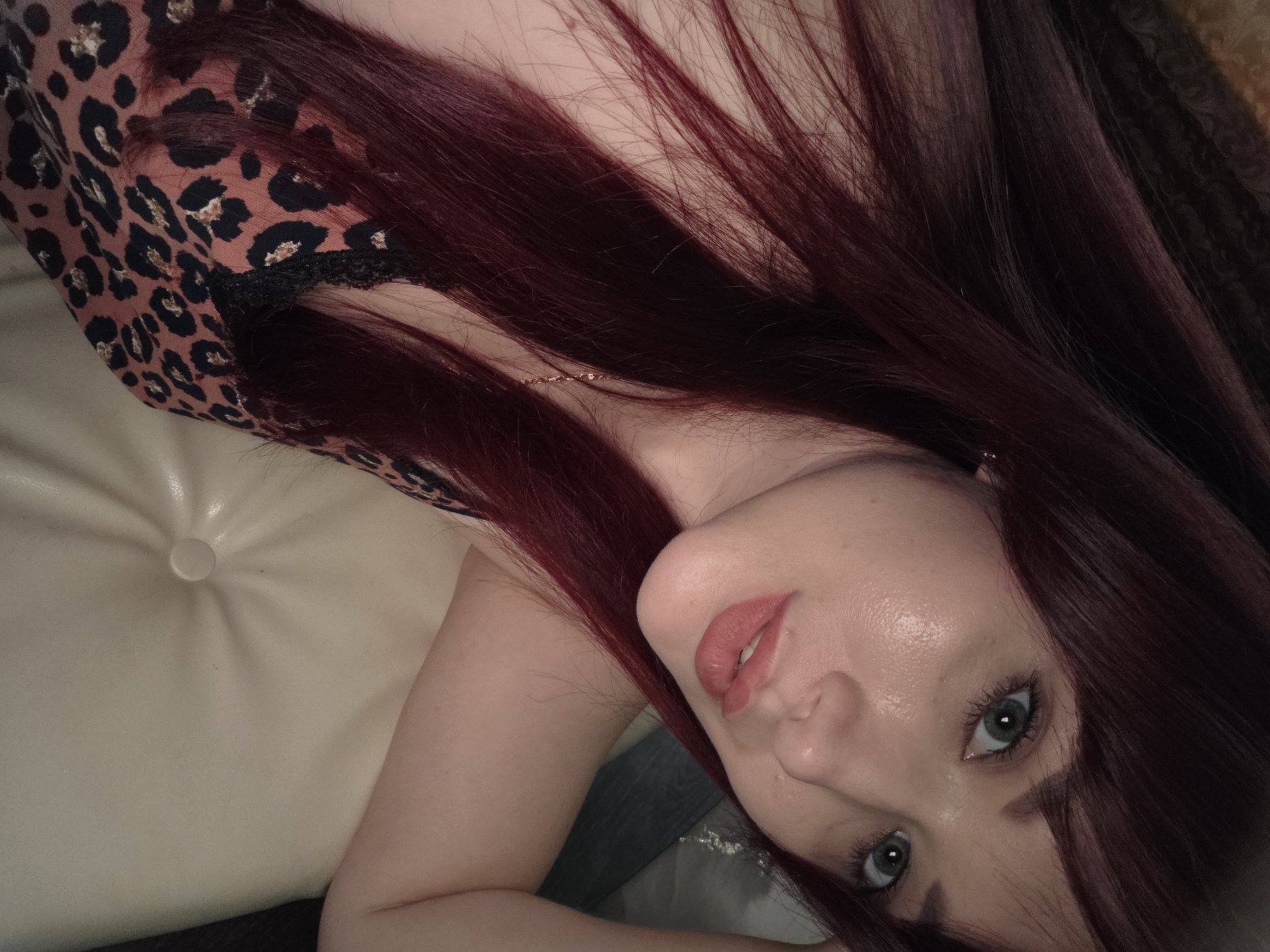 A close-up selfie of a woman with long, dark red hair and green eyes, wearing a leopard print top with black lace trim. She is resting her head on her arm, which is on a light-colored, tufted surface. The lighting is dim, creating highlights on her forehead and cheekbones. The background is out of focus and appears to be a patterned fabric. The image does not provide any information about the location in Chaikovsky, Russia.