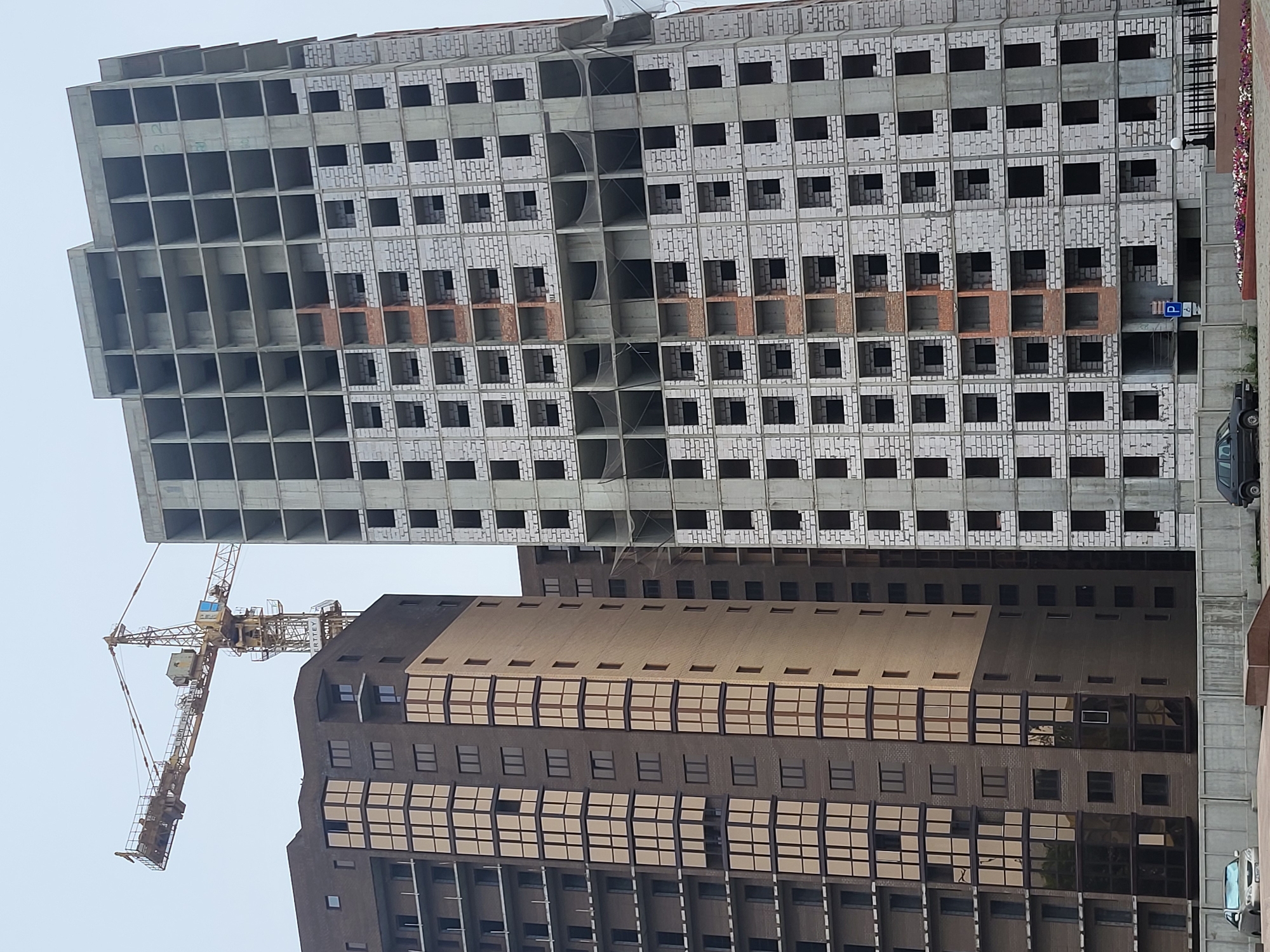 In Petropavl, Kazakhstan, a yellow tower crane is positioned in front of two under-construction high-rise buildings. The building on the left is clad in dark brown brick and beige panels, with numerous windows. The building on the right is made of concrete and white blocks, with empty window frames. The sky is overcast. In the foreground, a concrete fence separates the construction site from a street where two cars are parked.