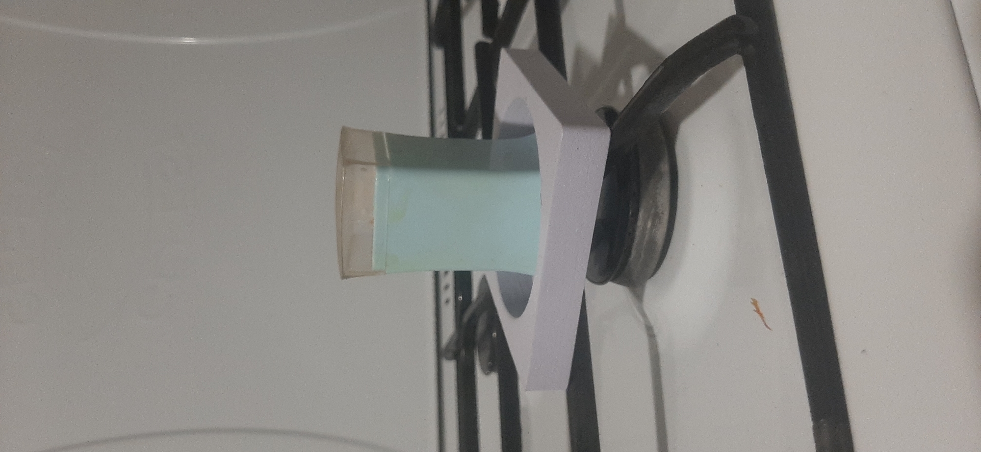 A light blue, rectangular object with a clear plastic top is placed inside a light purple, square holder. The holder is positioned on a gas stove burner. The background is a white stovetop and the interior of a stove oven.