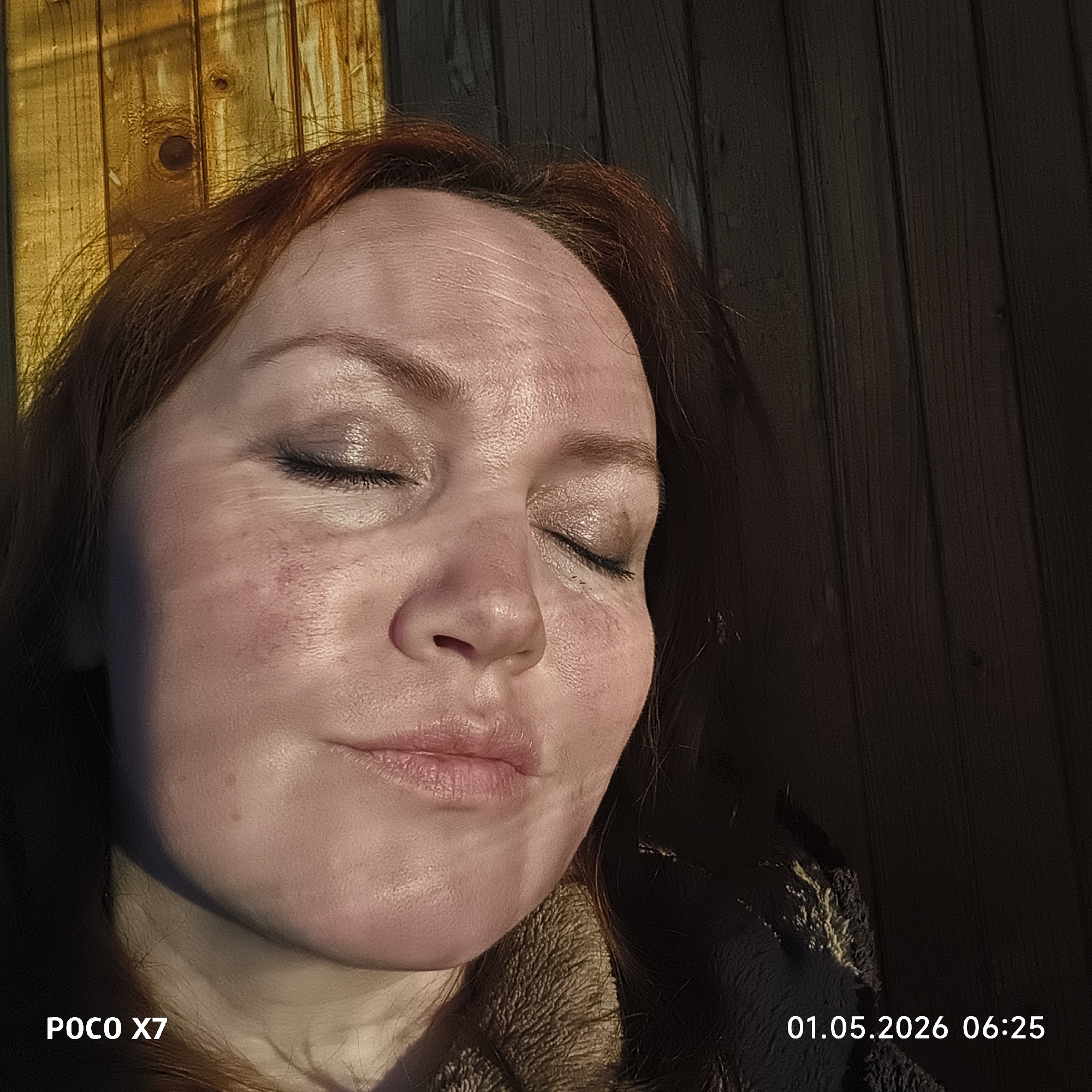 A close-up, portrait-style image features a woman with reddish-brown hair and fair skin, illuminated by strong, warm light, likely from the sun. Her eyes are closed, and her expression appears peaceful and relaxed, suggesting she is basking in the light. Her skin shows natural texture and some areas of redness, particularly on her cheeks and forehead. She is wearing a dark, textured garment with a fluffy or faux fur collar, visible around her neck and shoulder.
The background consists of a rustic wooden structure with vertical planks. The bright light source, positioned to the left, casts golden highlights on the wooden planks behind her head and the upper left of the frame, while the right side of the background is in deep shadow, revealing the wood grain.
In the bottom left corner of the image, the text "POCO X7" is visible, indicating the device used to capture the photo. The bottom right corner displays a timestamp "01.05.2026 06:25", suggesting the image was taken in the early morning, consistent with the quality of light. The overall scene conveys a tranquil moment, possibly outdoors, in Chaikovsky, Russia.