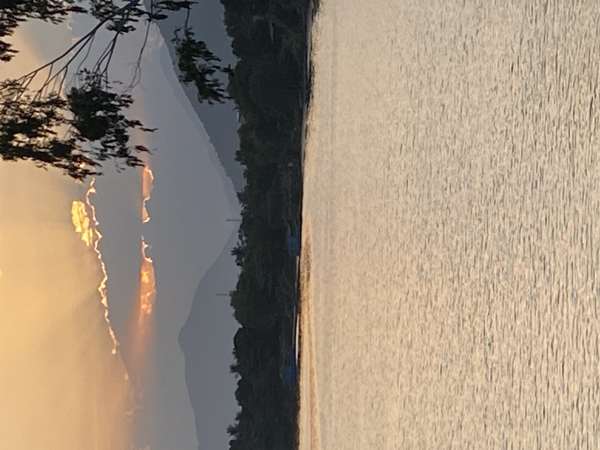 The image captures a serene sunset over a body of water, likely a river or bay, in Nha Trang, Vietnam. The sky is painted with warm hues of orange and yellow, with dramatic sun rays piercing through the clouds. In the foreground, the water reflects the sunset's glow, creating a shimmering effect. The middle ground features a lush green treeline bordering the water, with hints of structures and perhaps a few small boats. In the background, silhouetted mountains rise gently against the fading light. The overall atmosphere is one of tranquility and natural beauty.