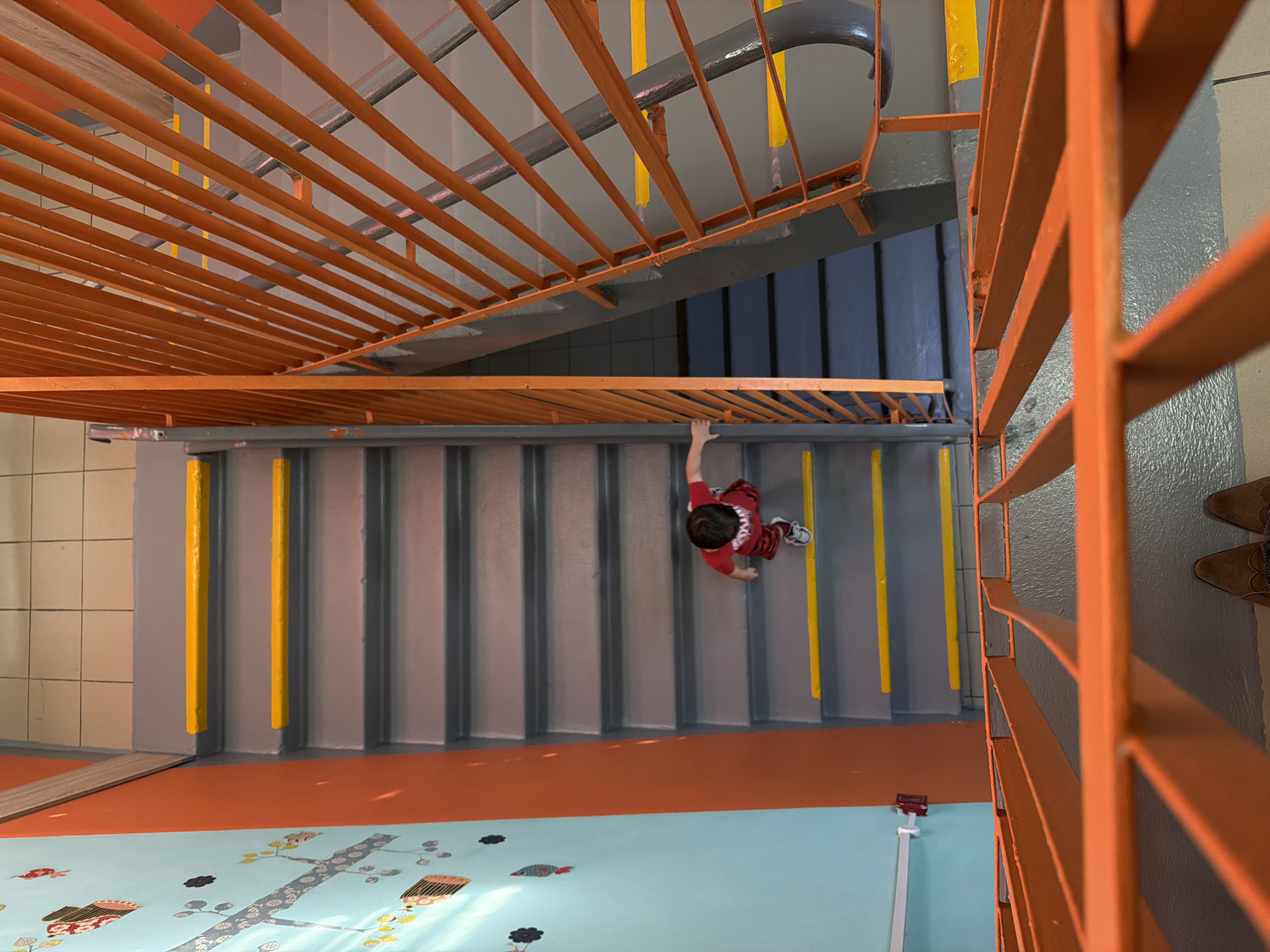 A high-angle shot looks down a flight of stairs in Khimki, Russia. The stairs are grey with yellow stripes near the edges. A child in a red shirt and patterned pants is midway down the stairs, facing downwards with one hand on the railing. Orange metal railings are visible on both sides of the stairs. Below the stairs, the floor is a combination of orange and light blue, with a graphic pattern of trees and flowers on the blue section. In the top right corner, part of a person's brown shoes is visible. The lighting is bright and diffused.