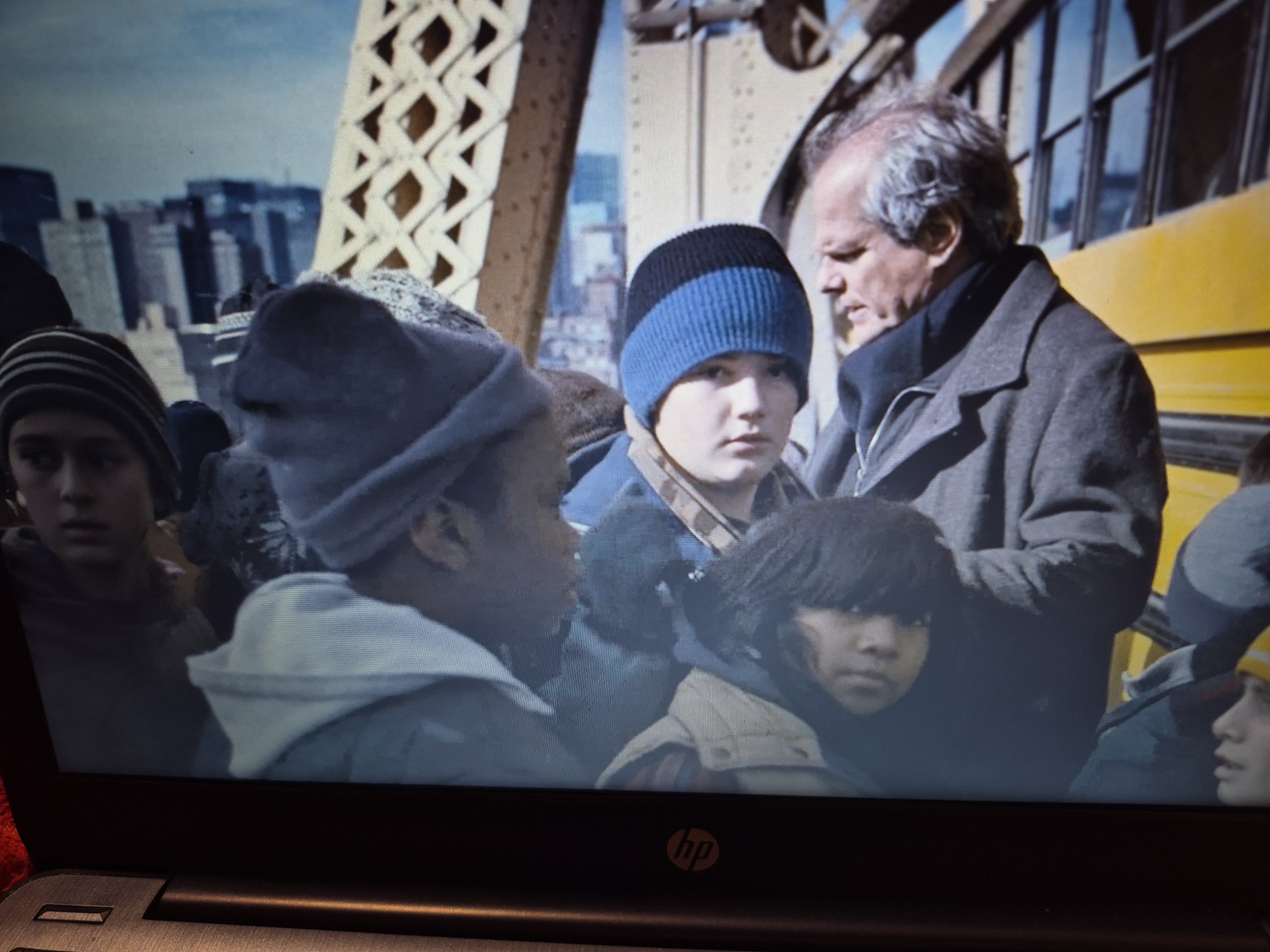 The image depicts a group of people, some of whom are wearing hats and coats, suggesting cold weather. A prominent yellow structure, possibly a bus, is visible in the background, along with a bridge with intricate lattice work and distant city buildings. The scene appears to be outdoors in an urban environment, with a focus on the faces and upper bodies of several individuals. The overall impression is one of a candid moment captured, possibly during a trip or outing. The presence of the yellow bus and bridge hints at a possible setting near a transportation hub or a scenic urban location. There are no clear indications of the city of Sandwell in the United Kingdom.