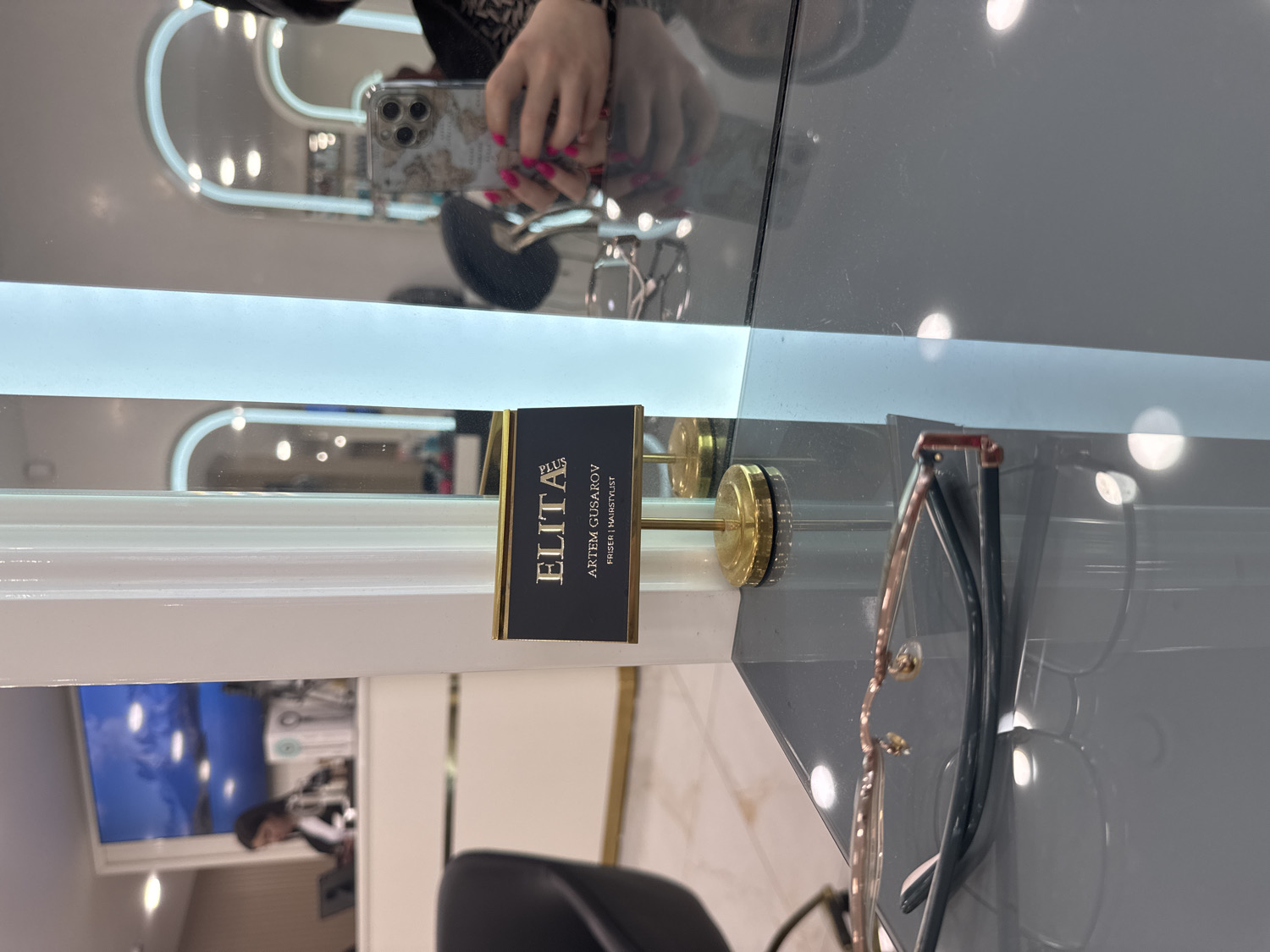 The image depicts an indoor scene within a modern commercial establishment, likely a hair salon or beauty parlor, possibly located in Sofia, Bulgaria. In the foreground, a pair of eyeglasses with rose gold frames and dark temples rests on a glossy, grey reflective surface.
Adjacent to the eyeglasses, a black and gold nameplate stands upright. The text "ELITA PLUS" is printed vertically along the left edge, while "ARTEM GUSAROV" and "FRISER | HAIRSTYLIST" are displayed horizontally. The nameplate is affixed to a golden, cylindrical stand.
The background is largely composed of reflections from a mirrored surface, possibly a wall or ceiling. A person's hands are prominently reflected, holding a smartphone with a decorative, light-colored case. The person's fingernails are painted bright pink. The smartphone's camera lenses are visible, suggesting it is actively being used to capture an image or video. Further reflections show arched, illuminated light fixtures, and a faint reflection of a monitor or television screen displaying a person can be seen in the lower left quadrant of the reflected area. The setting appears contemporary and well-lit, with polished and reflective surfaces contributing to a clean aesthetic.