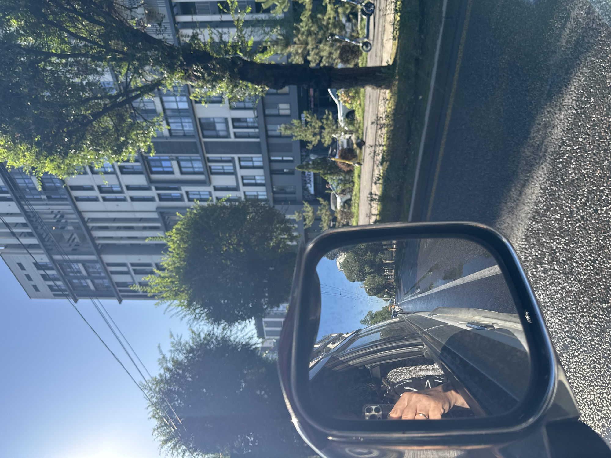 The image captures a street scene in Almaty, Kazakhstan, viewed from the passenger side mirror of a vehicle. The mirror reflects a road with wet patches, indicating recent rain, and a white lane marker. Trees line the roadside, and a modern, multi-story building with a grey and white facade stands in the background. The sky is clear and blue, with the sun shining brightly and creating lens flare. In the reflection, a person's hand, holding a smartphone, is visible through the car window, suggesting the photo was taken by someone inside the vehicle. On the street, there are two electric scooters parked and a white car. The pavement has a textured, speckled appearance. Overhead, power lines crisscross the sky.