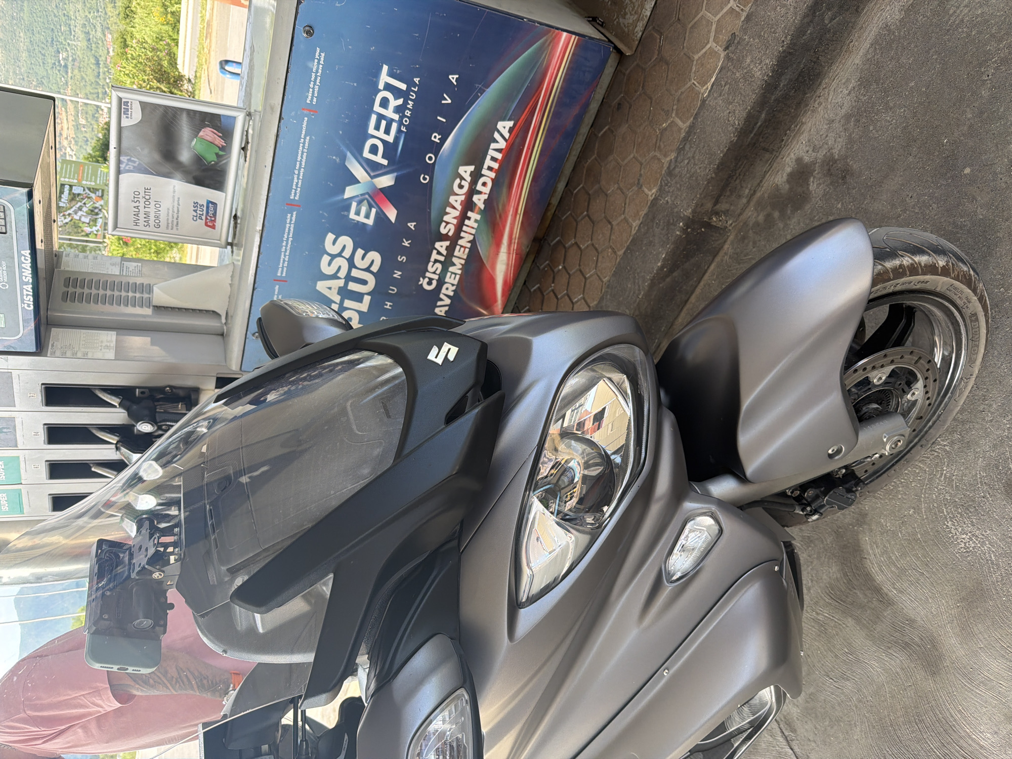 The image shows a gray Suzuki scooter parked at a gas station in Dub, Montenegro. The scooter is in the foreground, with its front end and windshield clearly visible. Behind the scooter, there are several gas pumps and a display board advertising fuel. A person wearing a pink shirt is partially visible in the background, standing near the pumps. The ground is paved with hexagonal tiles and concrete. The overall scene depicts a typical urban environment at a gas station.