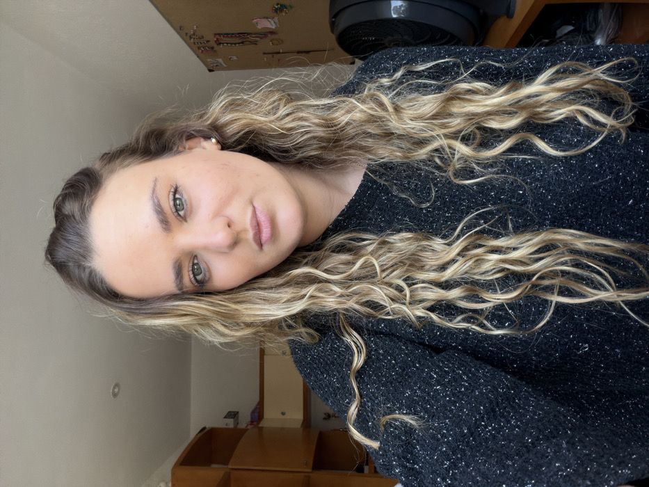 A fair-skinned woman with green eyes and wavy blonde hair wearing a dark, sparkly sweater poses at a slight angle. The background shows a wall with a few decorative items and a light-colored cabinet or shelf. A partially visible black fan sits atop a dark surface behind her head. Her face is the primary focus, capturing her relaxed expression and natural makeup.