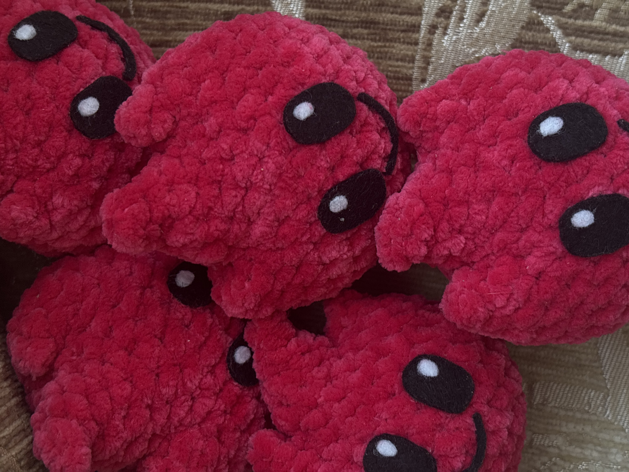 The image displays a close-up, overhead view of five small, plush, red, heart-shaped objects with black felt eyes and smiling mouths. These objects appear to be handmade, possibly crocheted or knitted, with a soft, fuzzy texture. They are arranged in a cluster on a textured, neutral-colored fabric background, which appears to be a woven material. No specific location in Varna, Bulgaria, is identifiable from the image.