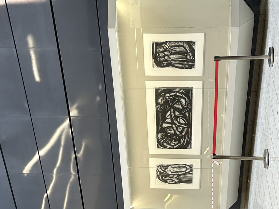 This media file captures an indoor public art display in what appears to be a modern setting, possibly a gallery, museum, or a public transit area in Nikea, Greece.
The central focus is a transparent display case showcasing three framed black and white prints. These artworks, likely woodcuts or linocuts, feature abstract or stylized figures with intertwining lines and forms. The largest print is positioned in the middle, oriented vertically, while the top and bottom prints are horizontally oriented. The artistic style is bold and graphic, using strong contrasts between black and white.
To the right of the display case, a polished metal stanchion with a red velvet rope serves as a barrier, indicating a designated viewing area or protection for the art. The floor is covered with light-colored, possibly tiled, surfaces.
On the left side of the image, a dark, reflective paneled wall dominates, showing dynamic, elongated streaks of bright light reflecting off its surface. These light patterns suggest ambient lighting or perhaps sunlight filtering in from outside. Faint, mirrored and upside-down text, possibly "KEA" or "23A", is barely discernible in the reflection on the display case, but too indistinct to confirm.
The scene appears to be well-lit, highlighting the artworks. There are no people visible, suggesting a quiet moment of observation. The clean lines and contemporary materials contribute to a sleek and sophisticated atmosphere.