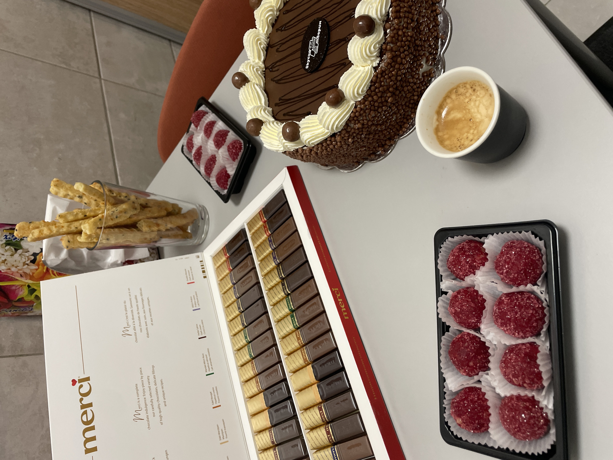 The image depicts a table laden with various treats, including a chocolate cake decorated with cream and chocolate balls, a box of Merci chocolates, a cup of coffee, a glass of breadsticks, and two small rectangular trays, one filled with what appears to be raspberry jellies and the other with smaller white and red candies. The objects are arranged on a light-colored tabletop. A corner of an orange chair is visible in the background, along with a beige wall.
Given that the cake contains the inscription "Вкусно творим" which translates to "Deliciously we create," combined with the Russian text written on the cake’s emblem, the setting could be a Russian or Eastern European context. However, there is no direct evidence within the image itself to confirm its exact location as Sofia, Bulgaria.