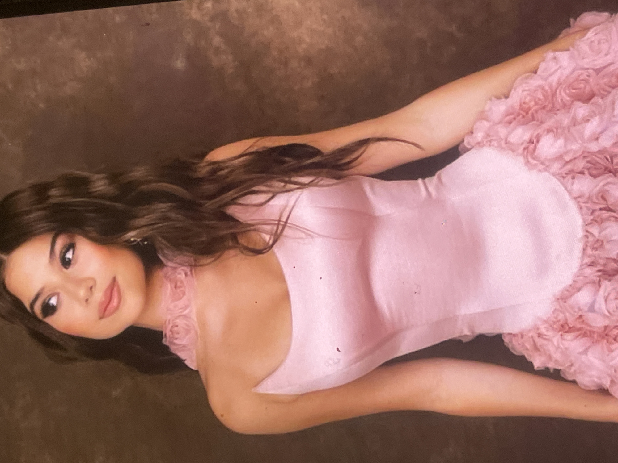 A portrait photograph captures a young woman posing gracefully, likely indoors in Poltava, Ukraine. She is lying on her back, with her head turned to the left, looking calmly towards the viewer. Her long, dark, wavy hair flows around her head and shoulders.
She is wearing an elegant, light pink gown. The dress features a smooth, fitted, strapless bodice, while the lower portion, from the waist down, is voluminous and adorned with intricate ruffles or fabric rosettes, giving it a textured, floral appearance. A matching ruffled detail is visible around her left shoulder and neck.
Her face is softly made up, with defined eyes and pink lips that complement her dress. The background is a mottled, dark brown, consistent with a studio backdrop. Soft, even lighting illuminates her features and the details of her attire, creating a serene and elegant mood. The image appears to be a photograph of a physical print or screen, as indicated by some minor specks. There is no visible text. The attire and setting suggest a formal occasion, such as a prom or a special event photoshoot.