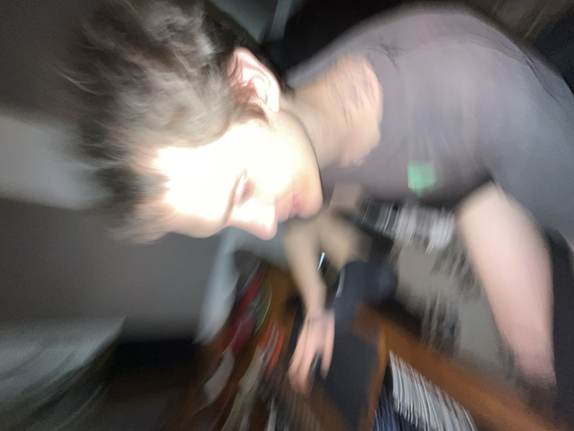 This media file presents a highly blurred and tilted image of a person, likely captured indoors. The significant motion blur throughout the image suggests either rapid movement of the subject or the camera during exposure.
The primary subject is a person whose head and upper torso are visible. They have light-colored, messy hair that appears to be swept back or wind-blown due to the blur. The left side of their face, including the nose, mouth, and chin, is heavily overexposed by a bright light source, making it difficult to discern specific facial features or expressions. The right ear is partially visible despite the blur.
The person is wearing a dark grey or black t-shirt, which features a faint, blurry green marking or logo on the chest. An arm and hand are visible towards the bottom right of the frame, appearing to hold an object, possibly a smartphone, although its exact form is obscured by blur.
The background is dark and indistinct, indicating an indoor setting. Hints of furniture, possibly a wooden surface, and various blurry objects with red, green, and white patterns are discernible in the lower-mid and lower-right sections, suggesting a potentially cluttered environment. The lighting is characterized by a very bright, direct source illuminating the subject's face, contrasting sharply with the dimness of the rest of the scene. No discernible text is present in the image. The time of day cannot be determined, but the lighting suggests an indoor setting with artificial light. The image provides no specific visual cues to its location in Vicente López, Argentina.