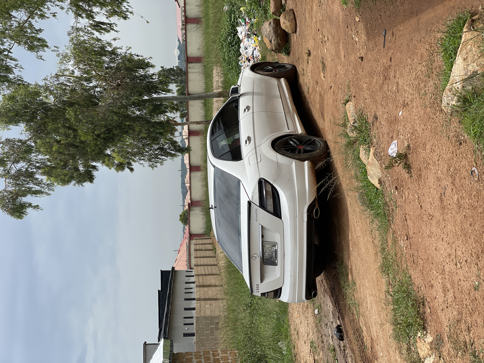 A white Mercedes-Benz C300 4MATIC sedan is parked on a dirt road, facing right, in Durumi, Nigeria. The car's rear is towards the camera, and it displays an AMG license plate frame. The vehicle is situated in a somewhat disheveled area, with overgrown grass and a pile of garbage visible to the right of the car. In the background, a low brick wall with red trim separates the immediate area from a street with houses and trees. The sky is overcast. The scene suggests an urban or suburban environment in Nigeria.