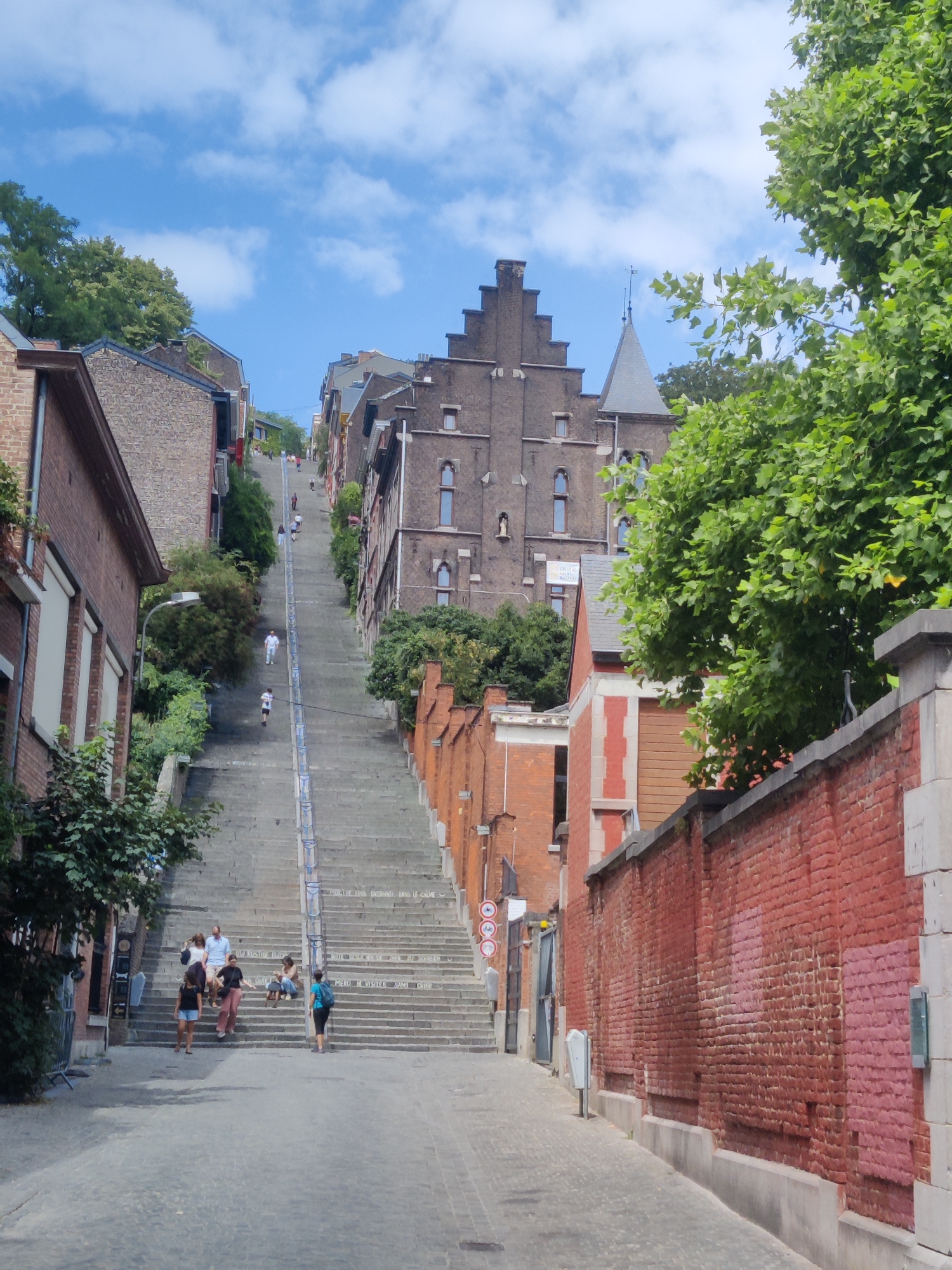 This image depicts the iconic Montagne de Bueren, a steep staircase in Liège, Belgium, featuring 374 steps. The perspective is from the bottom of the staircase, looking upwards. The sky above is a clear blue with scattered white clouds. Lush green trees line the sides of the staircase and the buildings, adding a vibrant contrast to the brick and stone architecture.
Several people are visible on the stairs, engaged in various activities. Some are ascending, while others are descending. In the foreground, a group of people are gathered on the lower steps, appearing to be taking a break or conversing. Further up the stairs, more individuals can be seen climbing.
The architecture of the buildings flanking the staircase is a mix of traditional brick construction with some stone elements. The building on the left in the foreground has a brick facade with large windows. The prominent building in the background, a historical structure with a stepped gable roof, dominates the upper portion of the image. The staircase itself is made of concrete or stone and has railings on either side. Along the sides of the staircase, there are also glimpses of residential buildings and the foliage of trees and bushes. The overall impression is a popular urban landmark with a lively atmosphere.