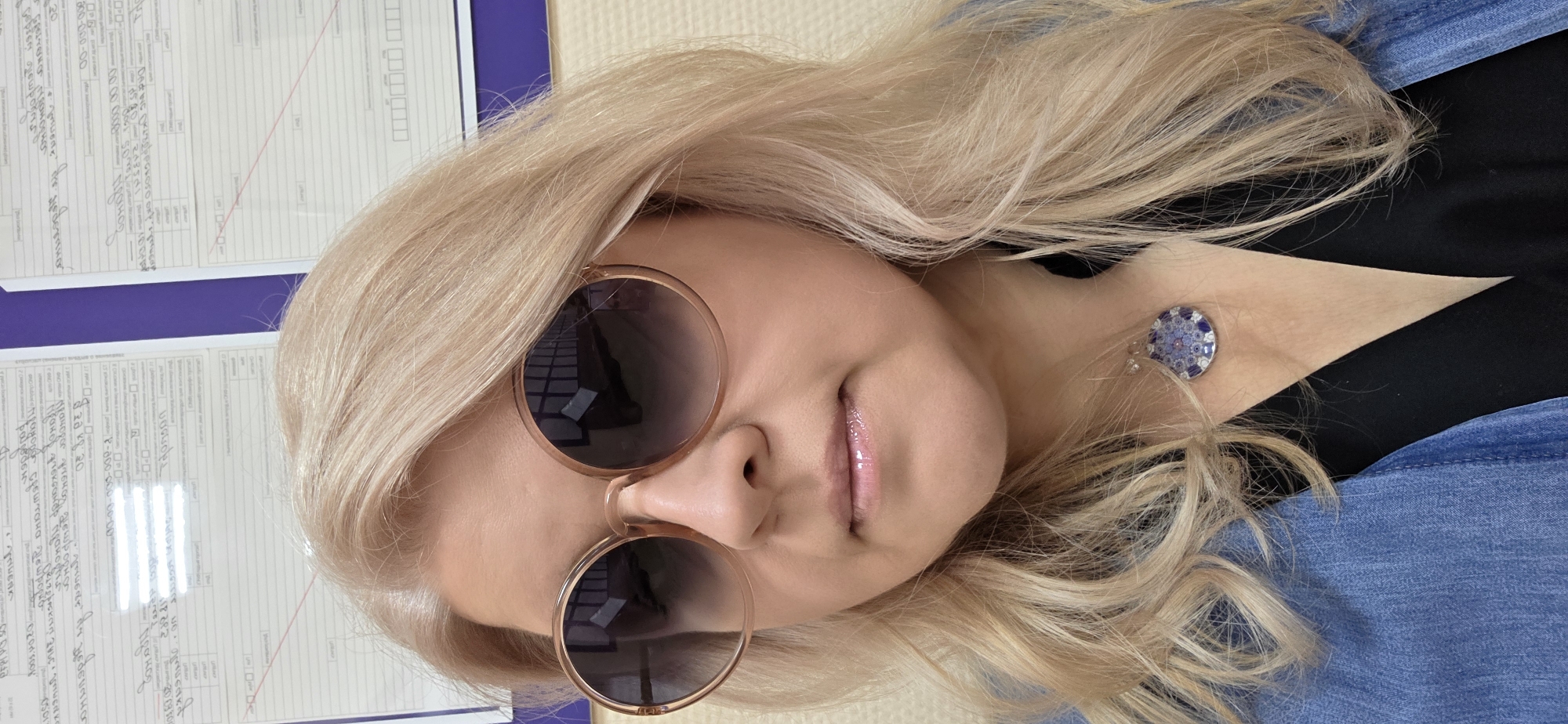 A tilted, close-up portrait features a woman with long, wavy blonde hair. She wears large, round, light brown-tinted sunglasses, which reflect indoor lighting. Her lips are glossy with a neutral expression. A distinctive circular piece of jewelry, possibly an earring or brooch, with intricate blue and white patterns, is visible on her neck/ear area. She is dressed in a black garment layered with a blue denim-like jacket or shirt.
The background shows several sheets of white paper with handwritten and printed text, affixed to a purple surface. This suggests an indoor setting, likely an office or administrative environment, with bright, possibly artificial, lighting.
Visible text on the papers, partially in Cyrillic, indicates form fields or documentation. The extracted text includes: "80-000-00", "20", "2009101", "00-00-000-000-2", "2048 20", "Праноз", "pp gmuser de you", "Пранодог учено дешродног прахода срεωτικα γισωρορμό", and "dr padevakor 0501".